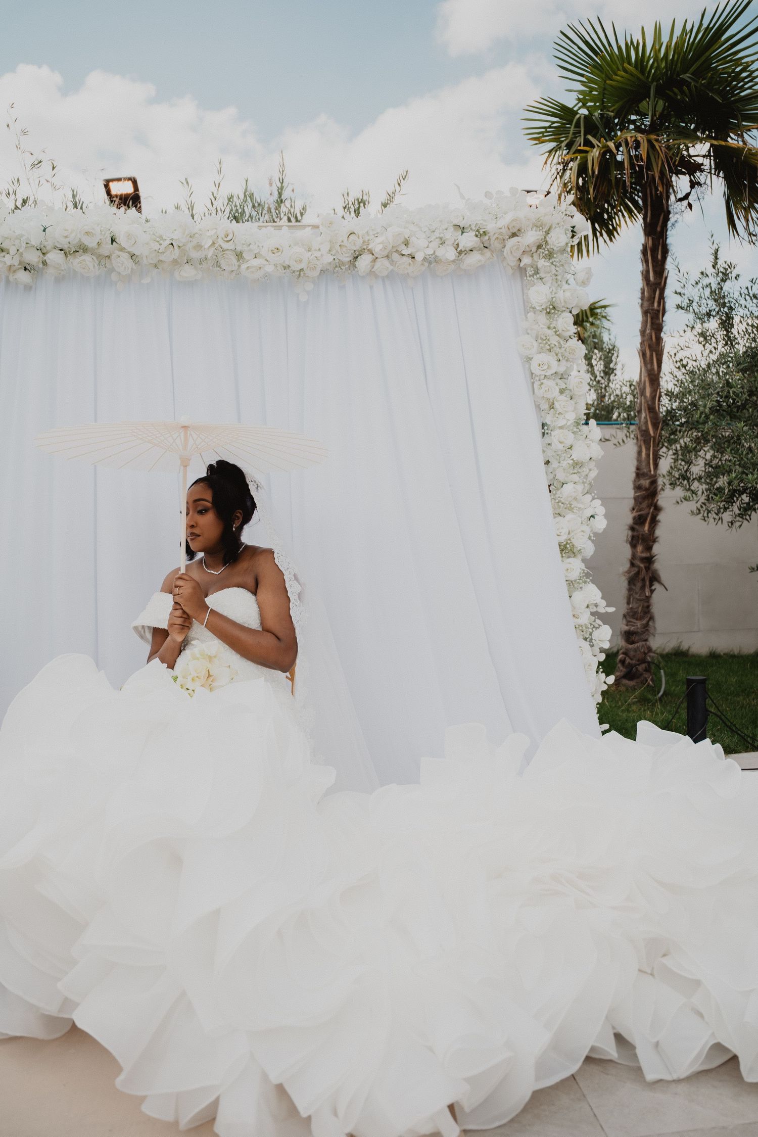 Une mariée vêtue d'une robe de mariée blanche volumineuse pose devant un fond blanc orné de palmiers.