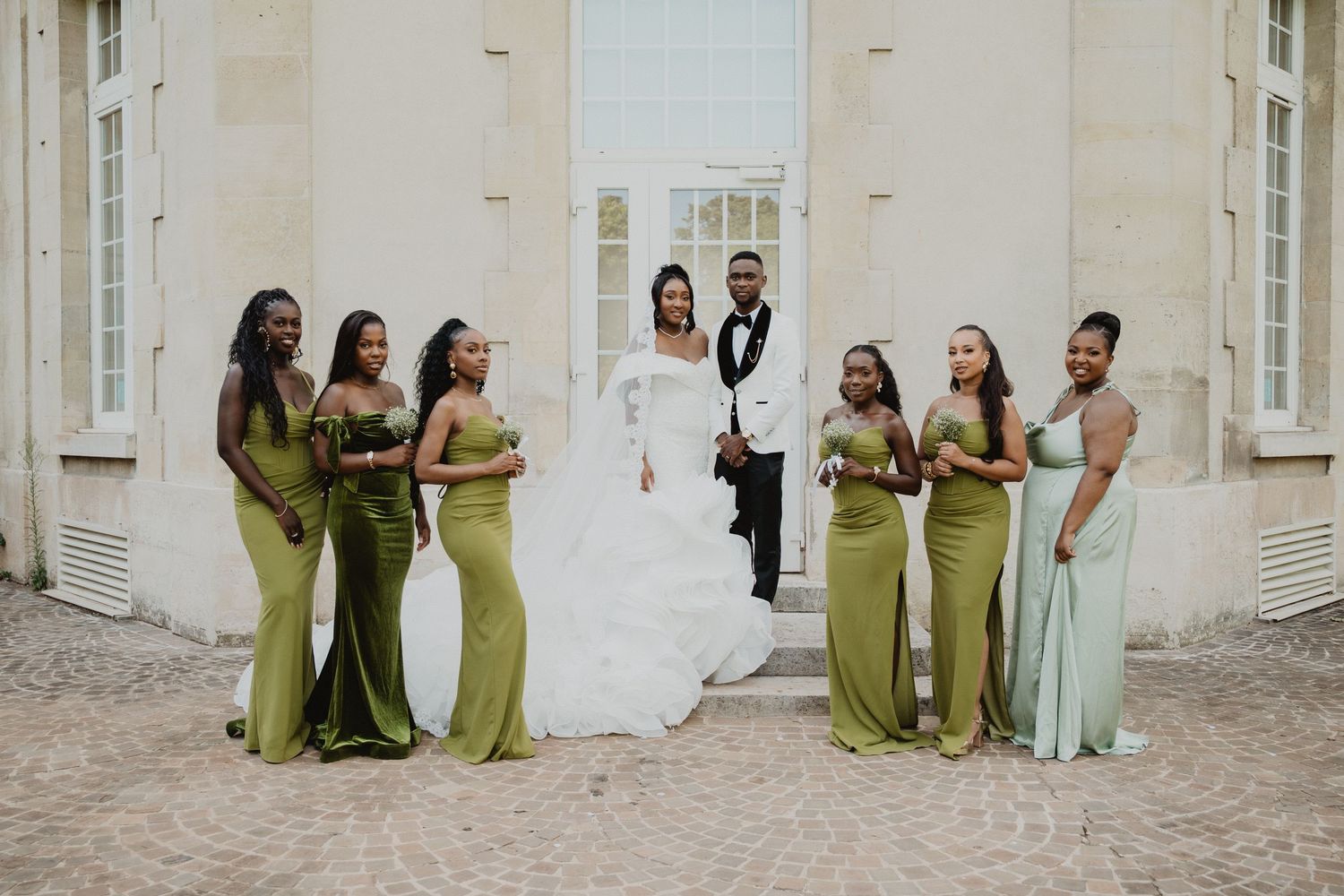 Les membres du cortège nuptial, vêtus de robes vert olive, posent devant la façade blanche d'un bâtiment.