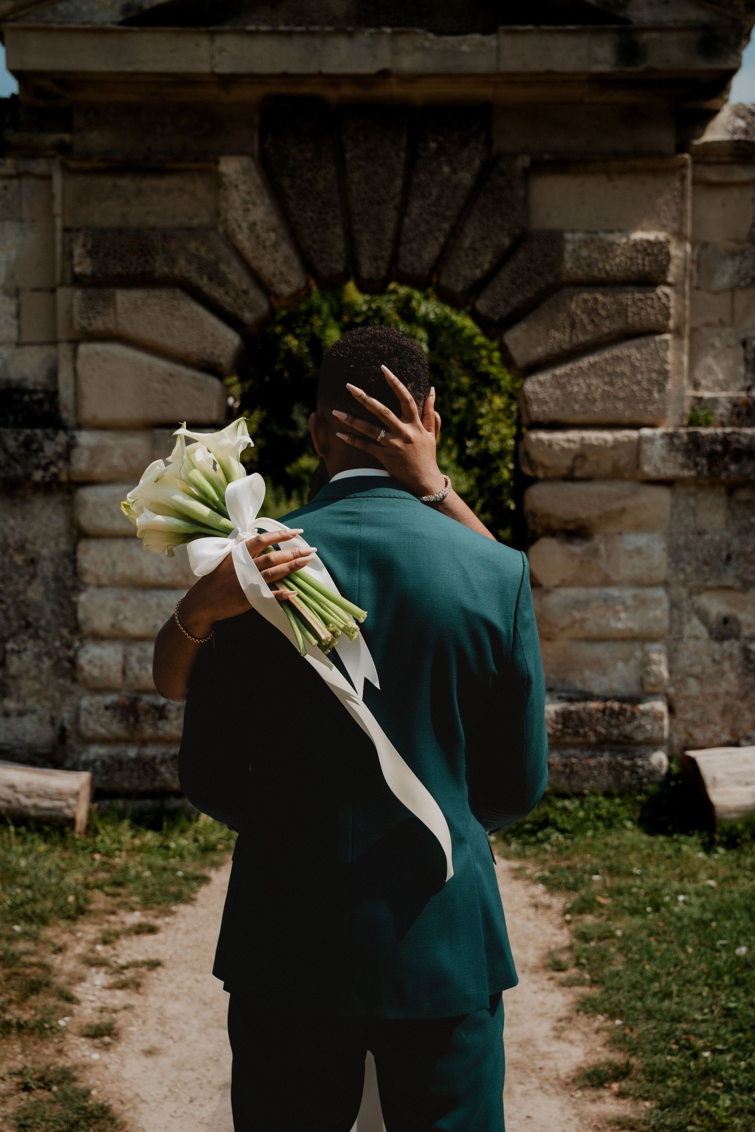 A couple embraces in front of a stone wall, with a white floral boutonniere visible.