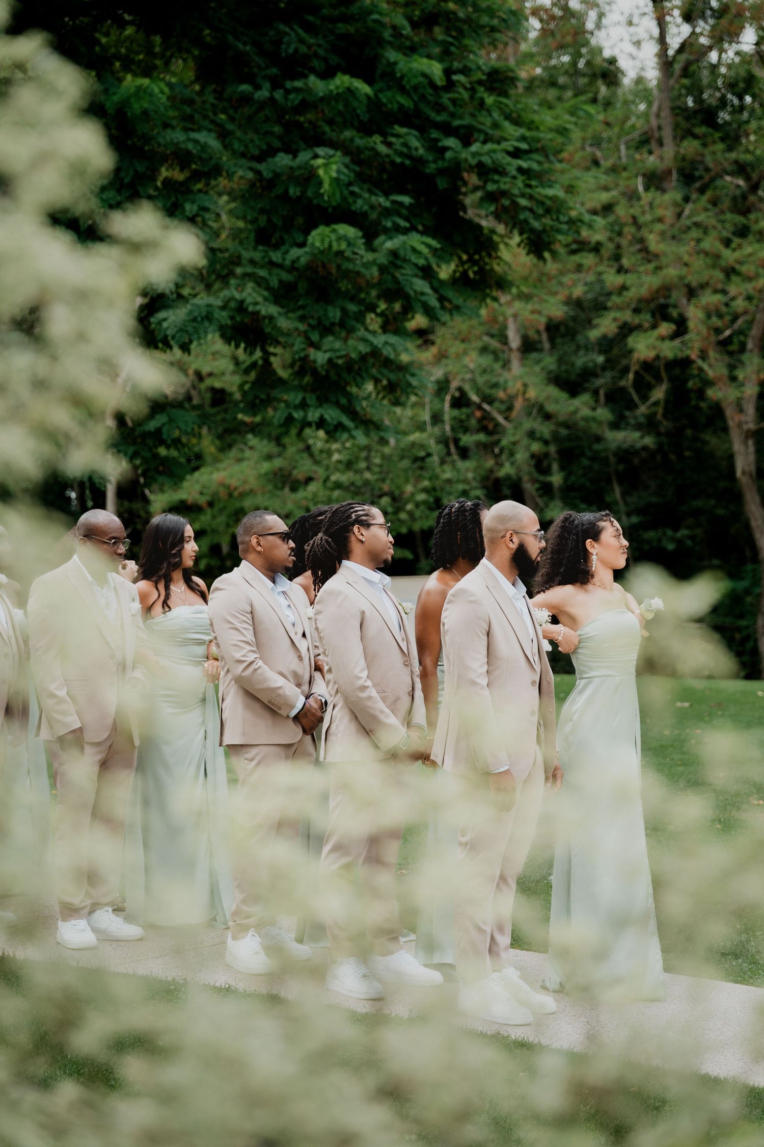 Wedding party in cream-colored attire poses outdoors among lush green foliage.