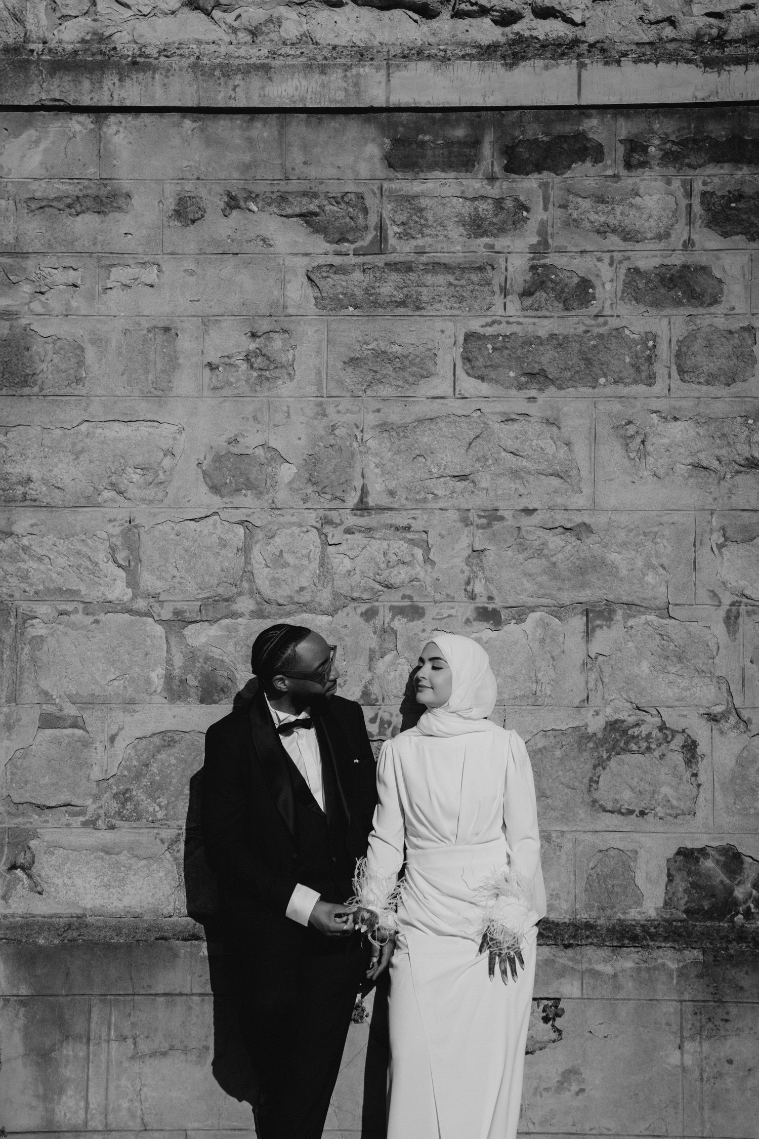 Black and white portrait of a couple in formal wear against a textured stone wall.