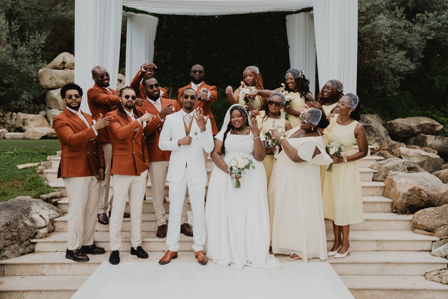 Wedding party poses together in coordinated orange and cream outfits under an outdoor gazebo.