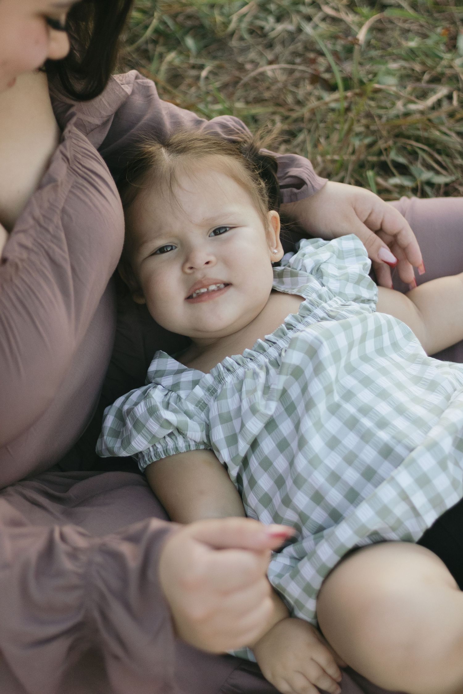 Adorable toddler girl in gingham dress lying in mothers lap smiling at camera outdoors.