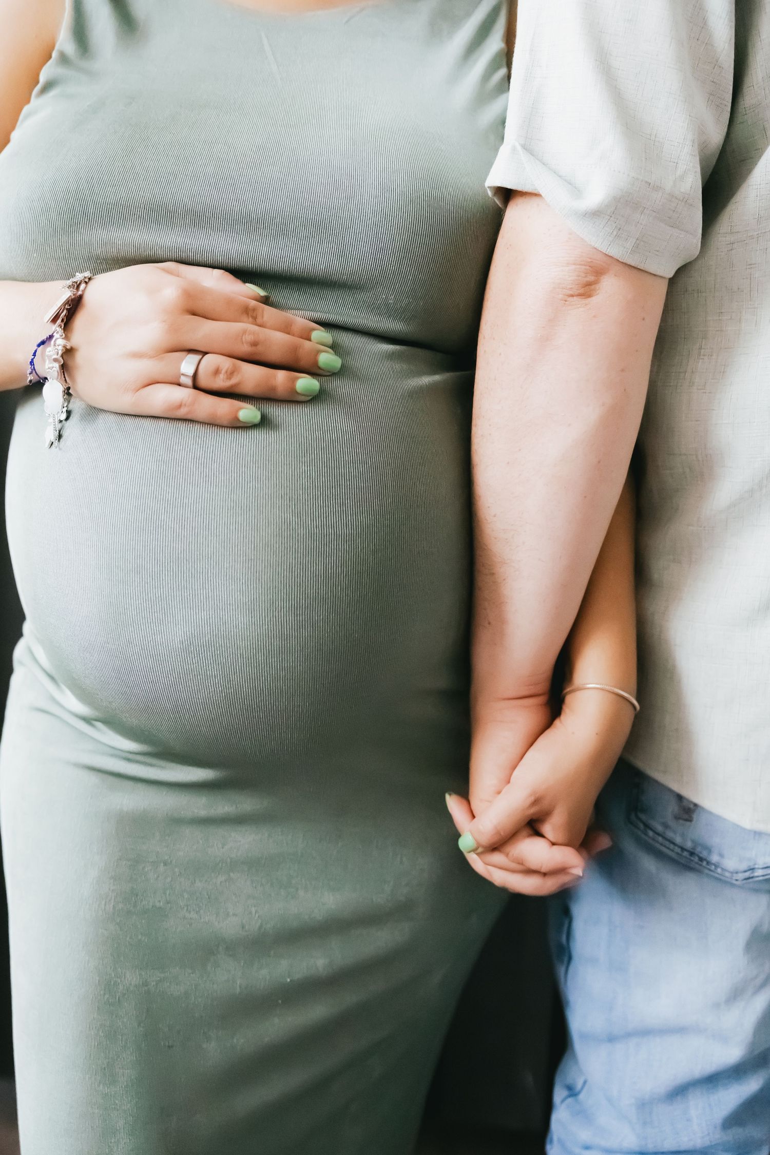A close-up of a pregnant woman in a sage green dress holding her baby bump while her partner holds her hand.