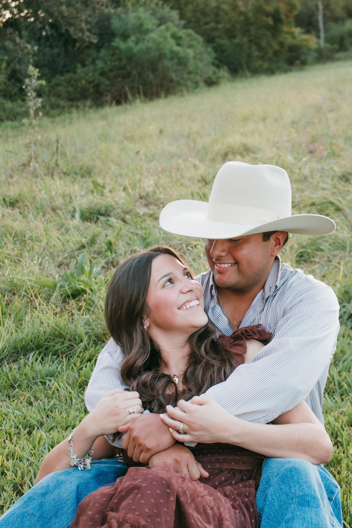 Couple sitting in green field, man in white cowboy hat embracing smiling woman in rustic outdoor engagement photo.