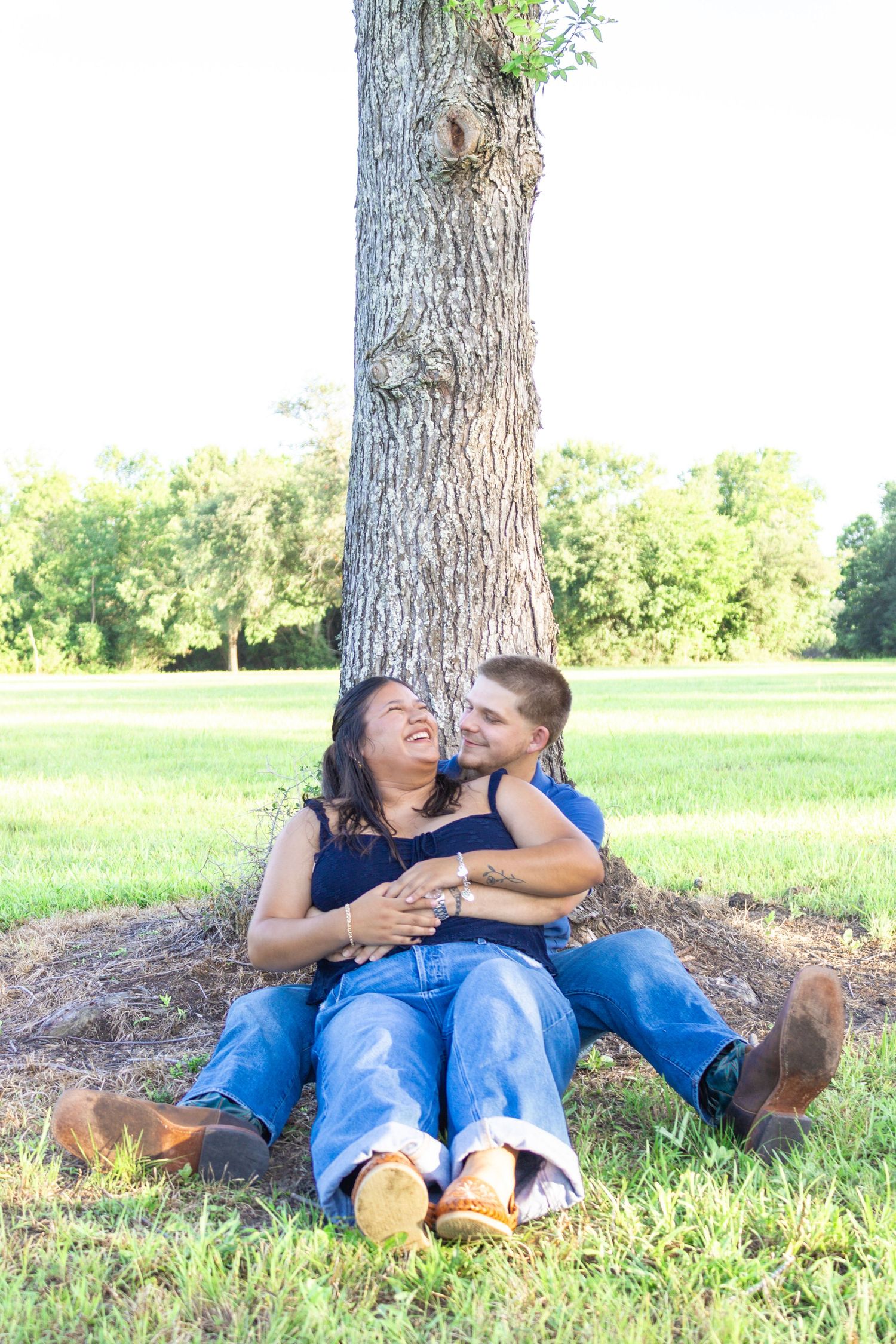 Happy couple laughing together while sitting against a tree trunk in a lush green park.