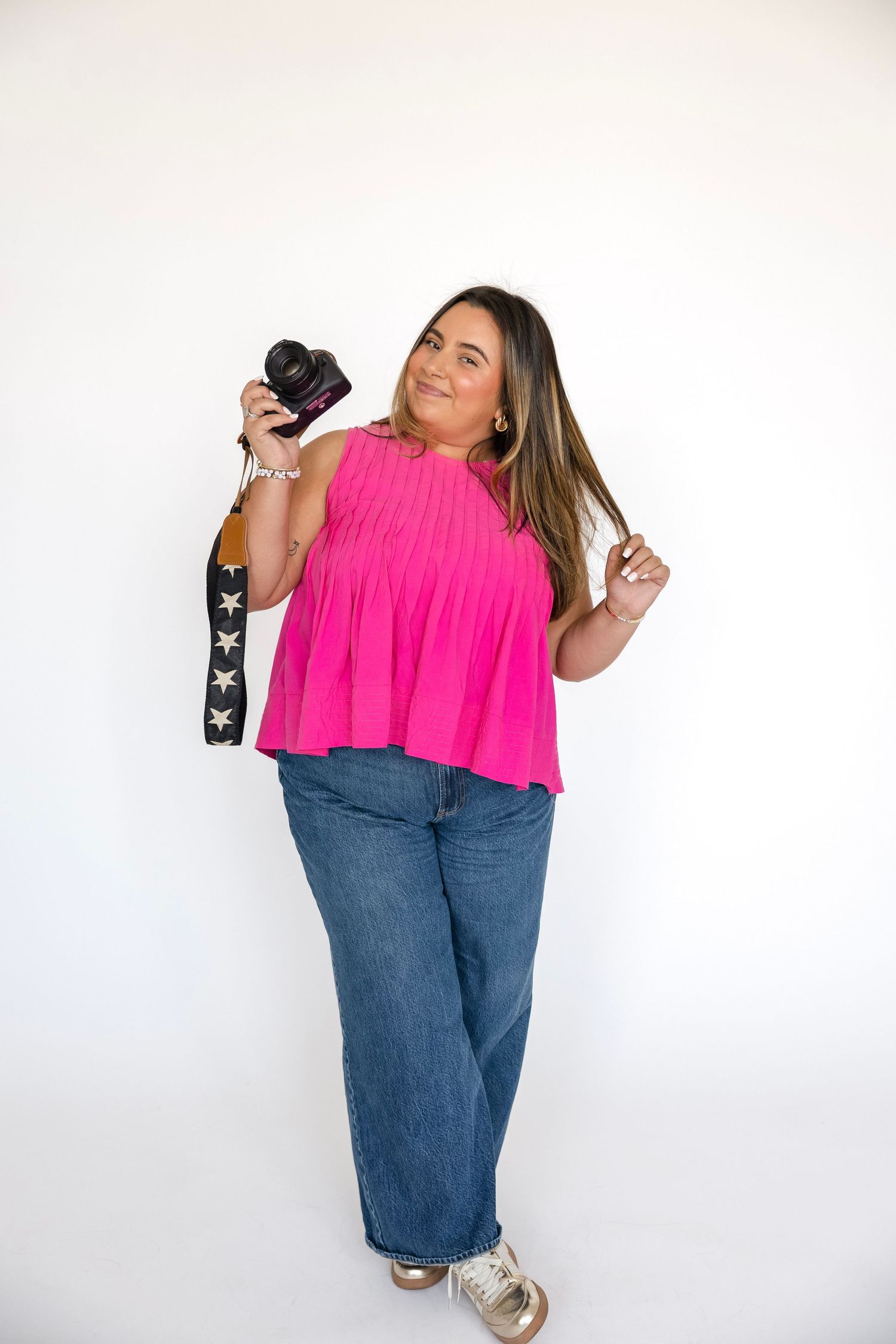 Cheerful woman in pink top holding a camera with star-print strap against white background.