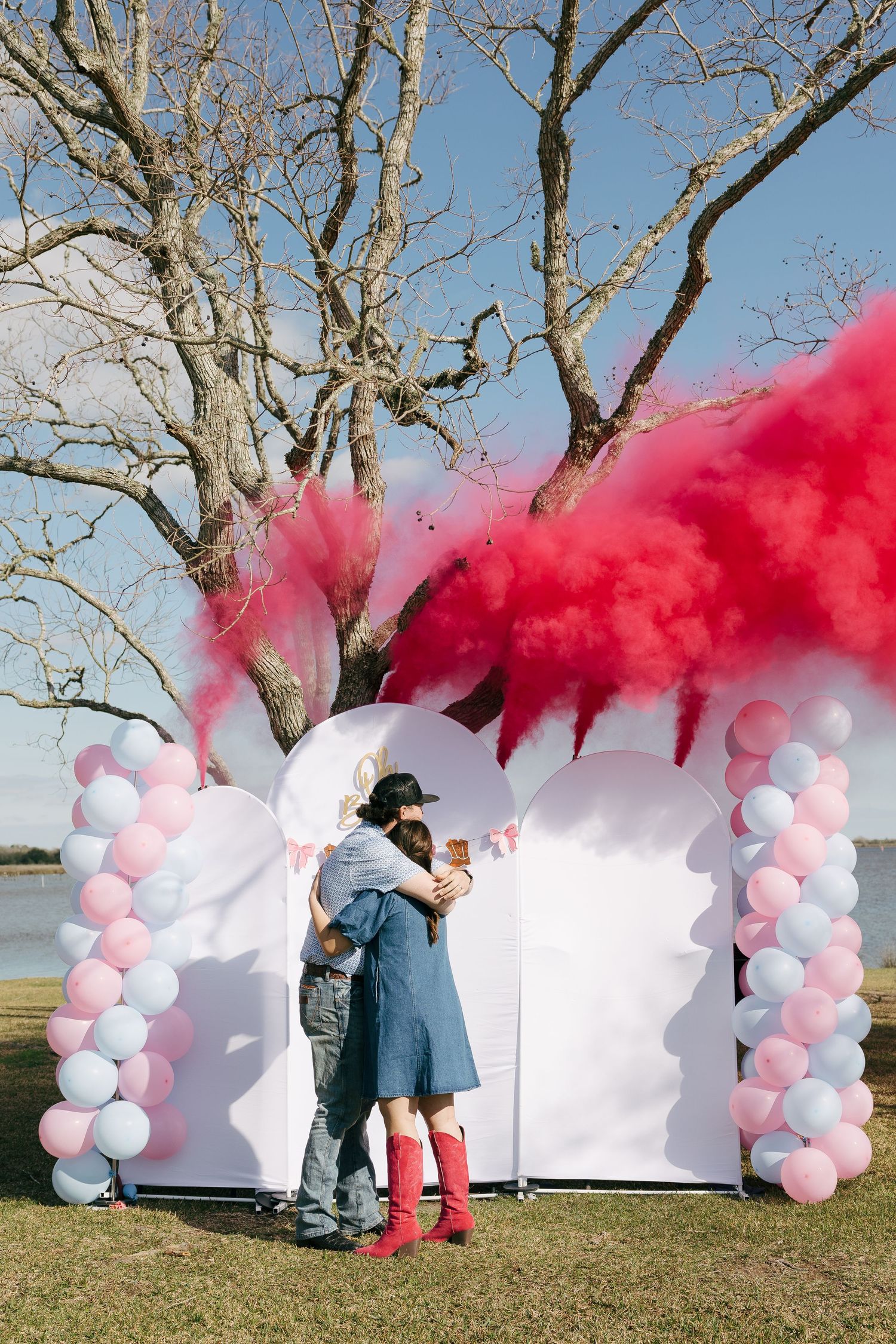 Couple embracing at gender reveal party with pink smoke explosion amid balloon arch by lakeside tree.