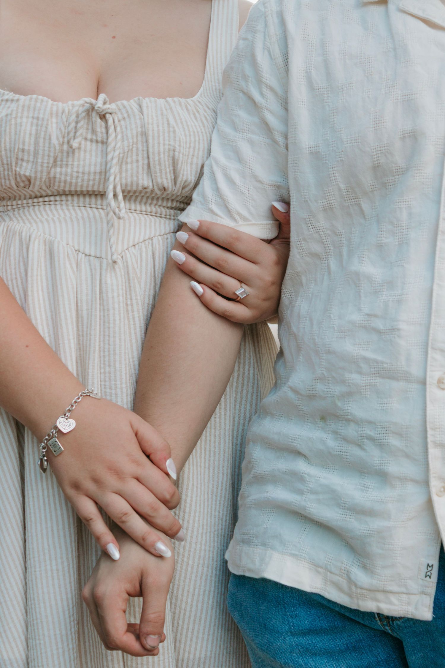 Close-up of couple holding arms showing engagement ring, diamond bracelet, and white manicured nails outdoors.