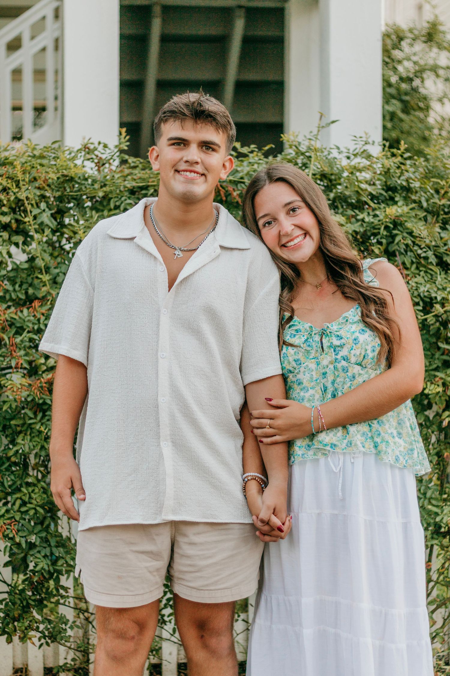 Smiling teenage couple posing together in front of green bushes, girl in floral dress leaning on boys shoulder.