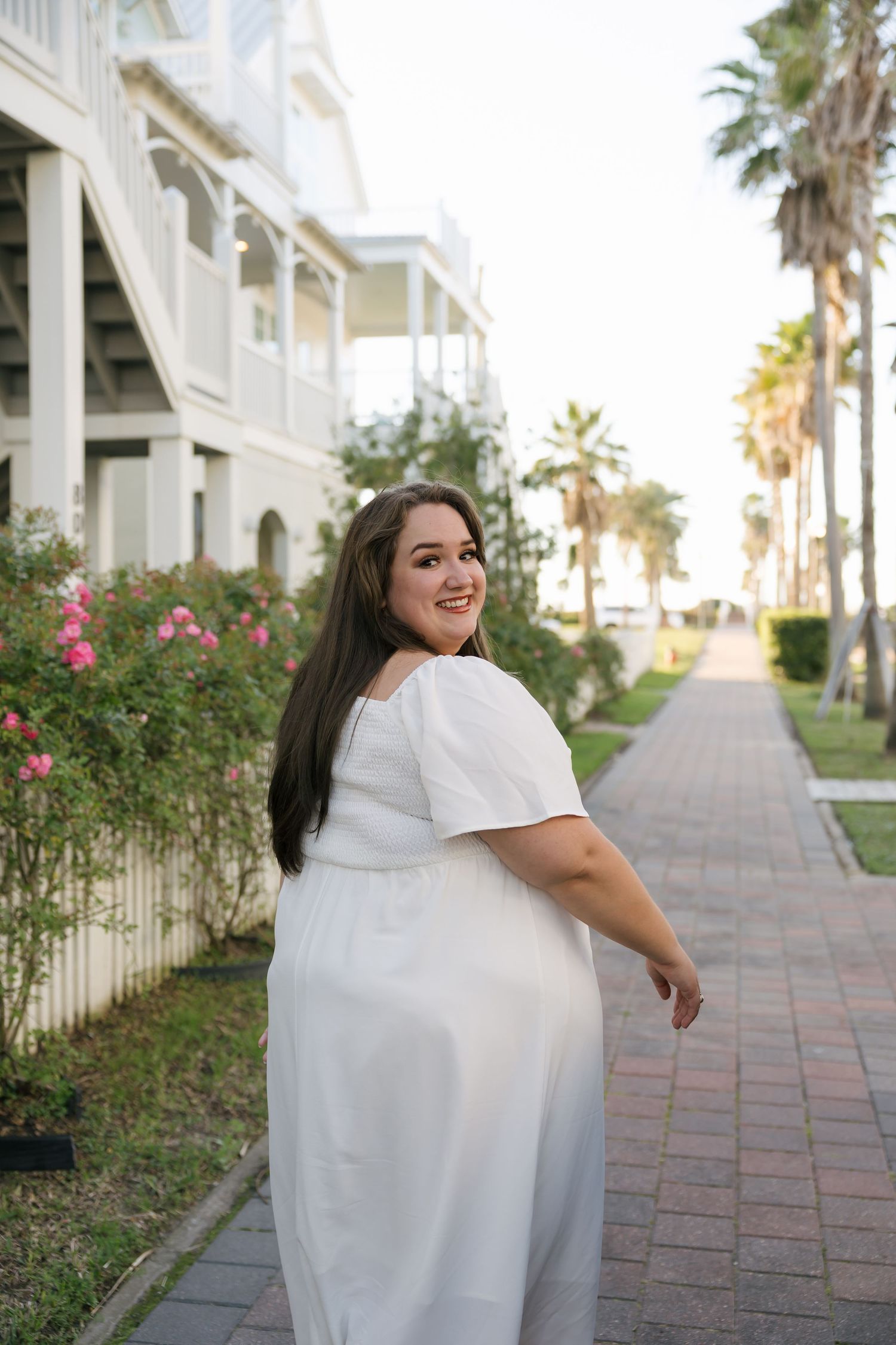 Smiling plus-size woman in white off-shoulder dress walking on brick path lined with palm trees and flowers.