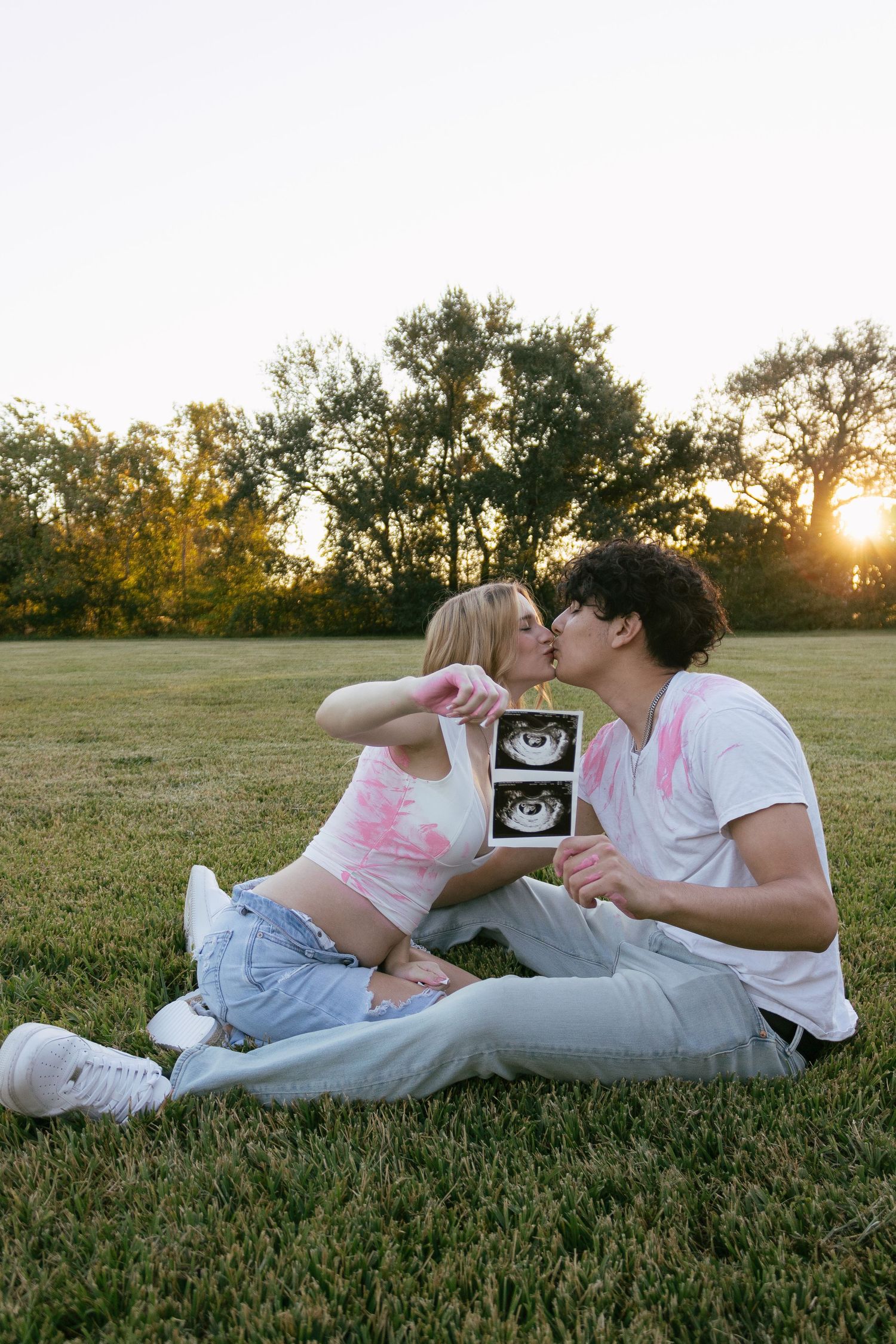 Couple sitting on grass kissing while holding ultrasound photo at sunset in a park.