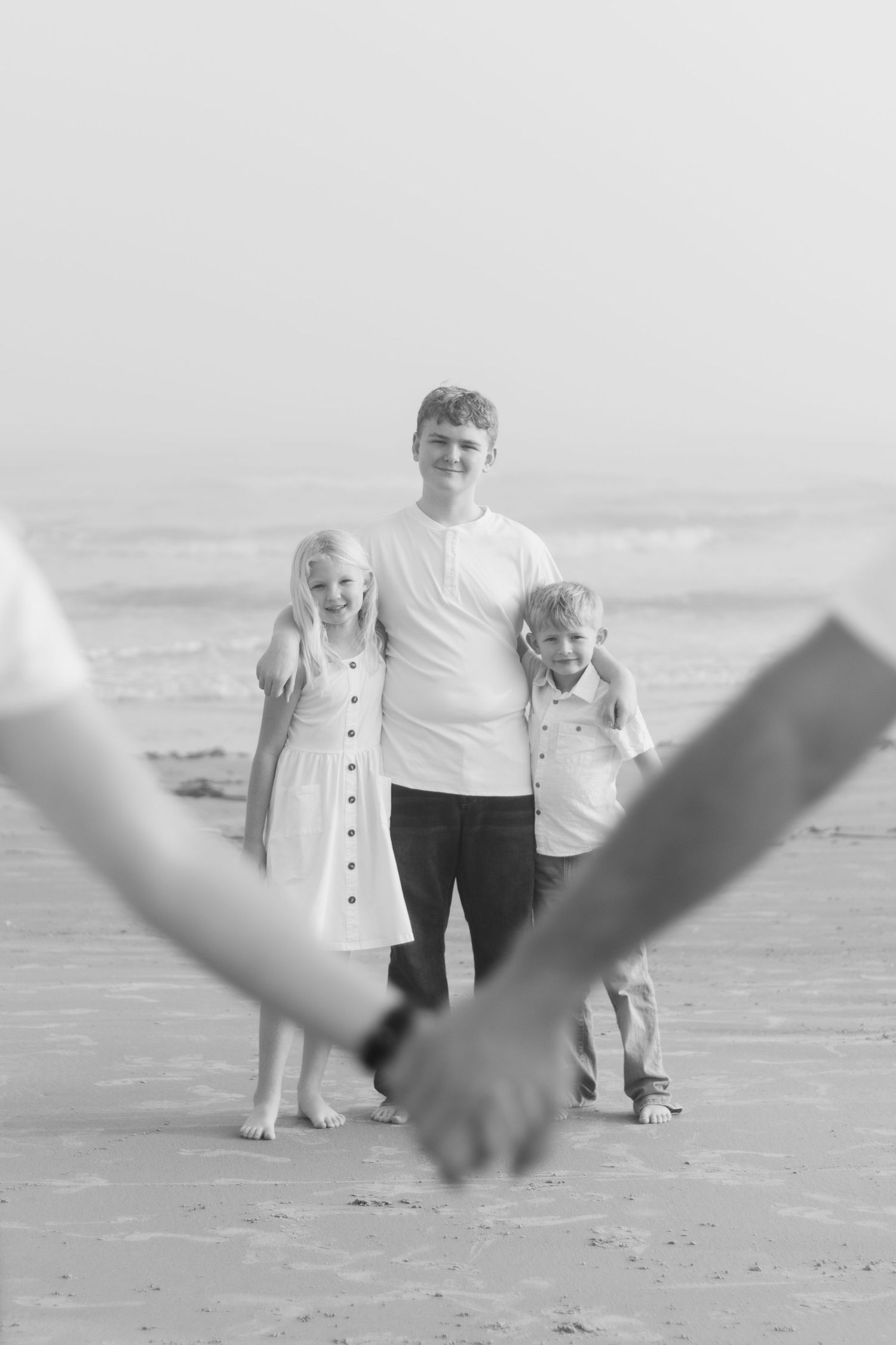 Black and white beach photo of three siblings posing while parents hold hands in foreground.