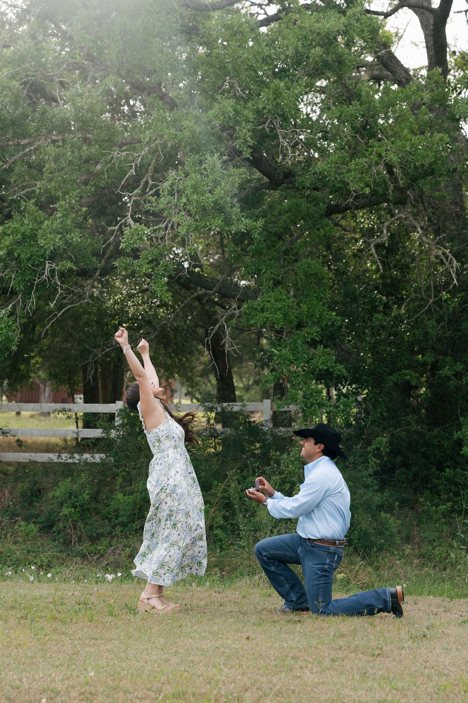 Man on one knee proposing to a woman with arms raised joyfully in a lush green yard.