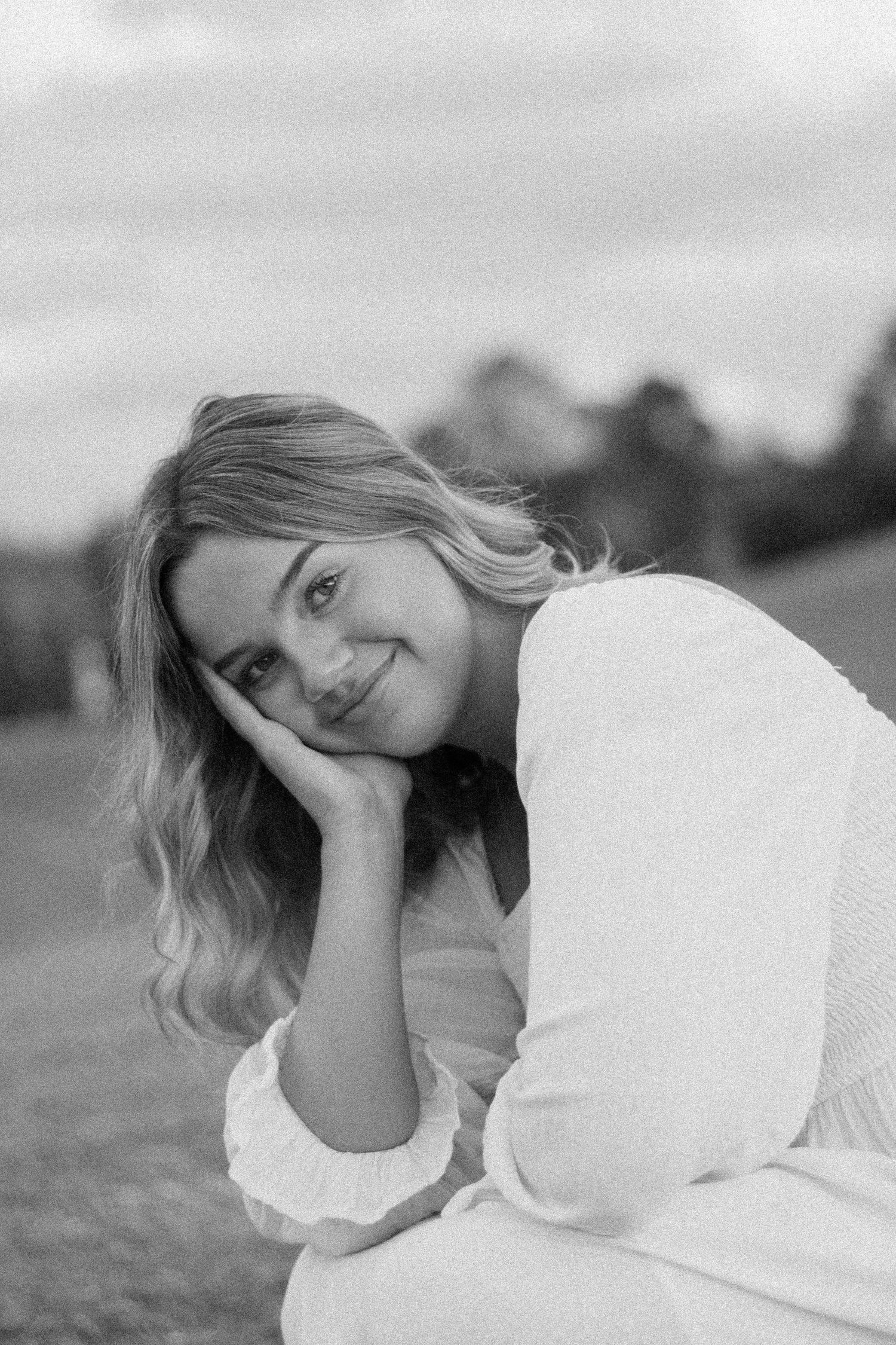 Black and white portrait of smiling blonde woman resting head on hand in outdoor setting.