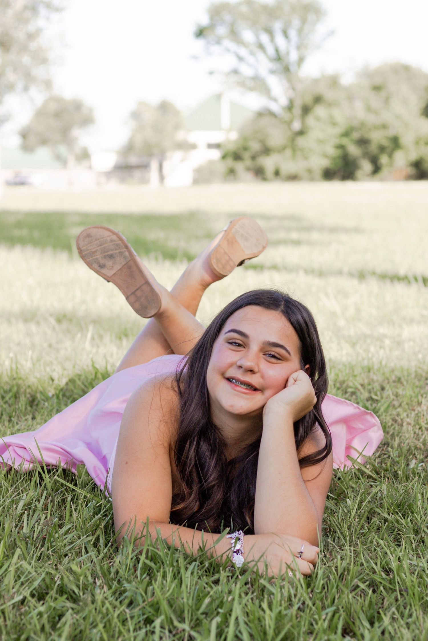Smiling teenage girl in pink dress lying on green grass in a sunny park looking at camera.