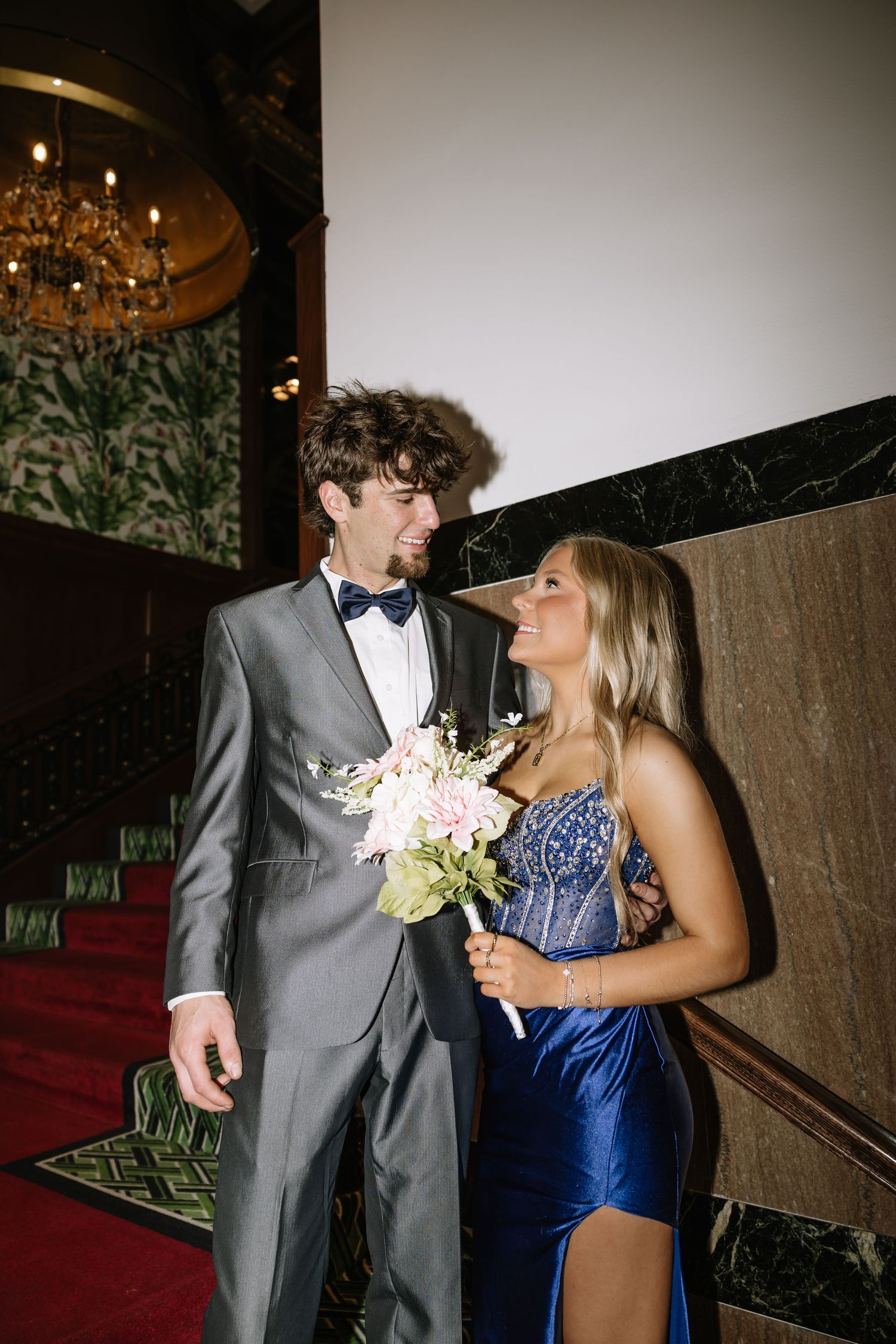 Couple in formal attire holding a bouquet smiling at each other in an elegant venue.
