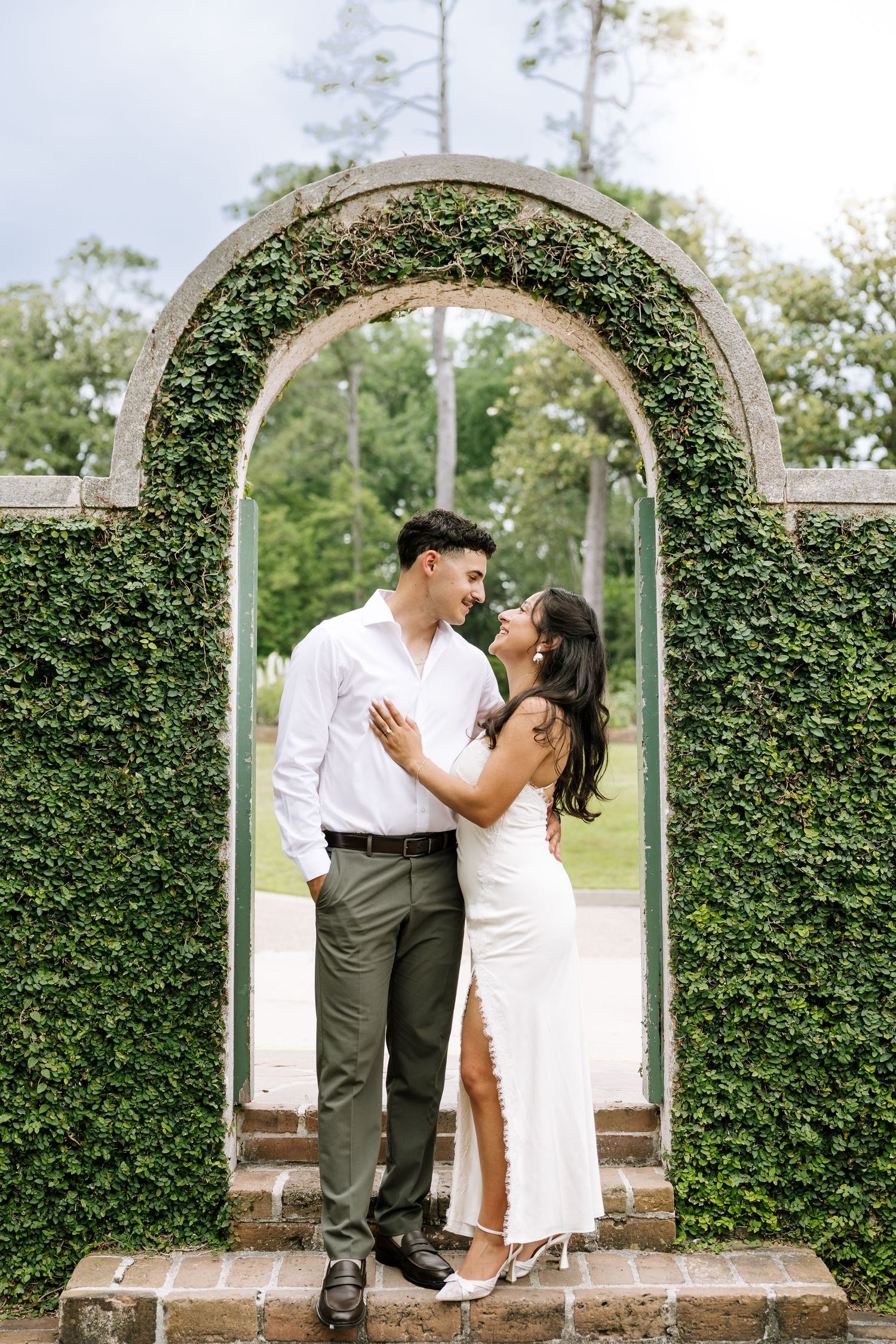 Couple almost kissing under an ivy-covered stone arch in a lush garden setting.