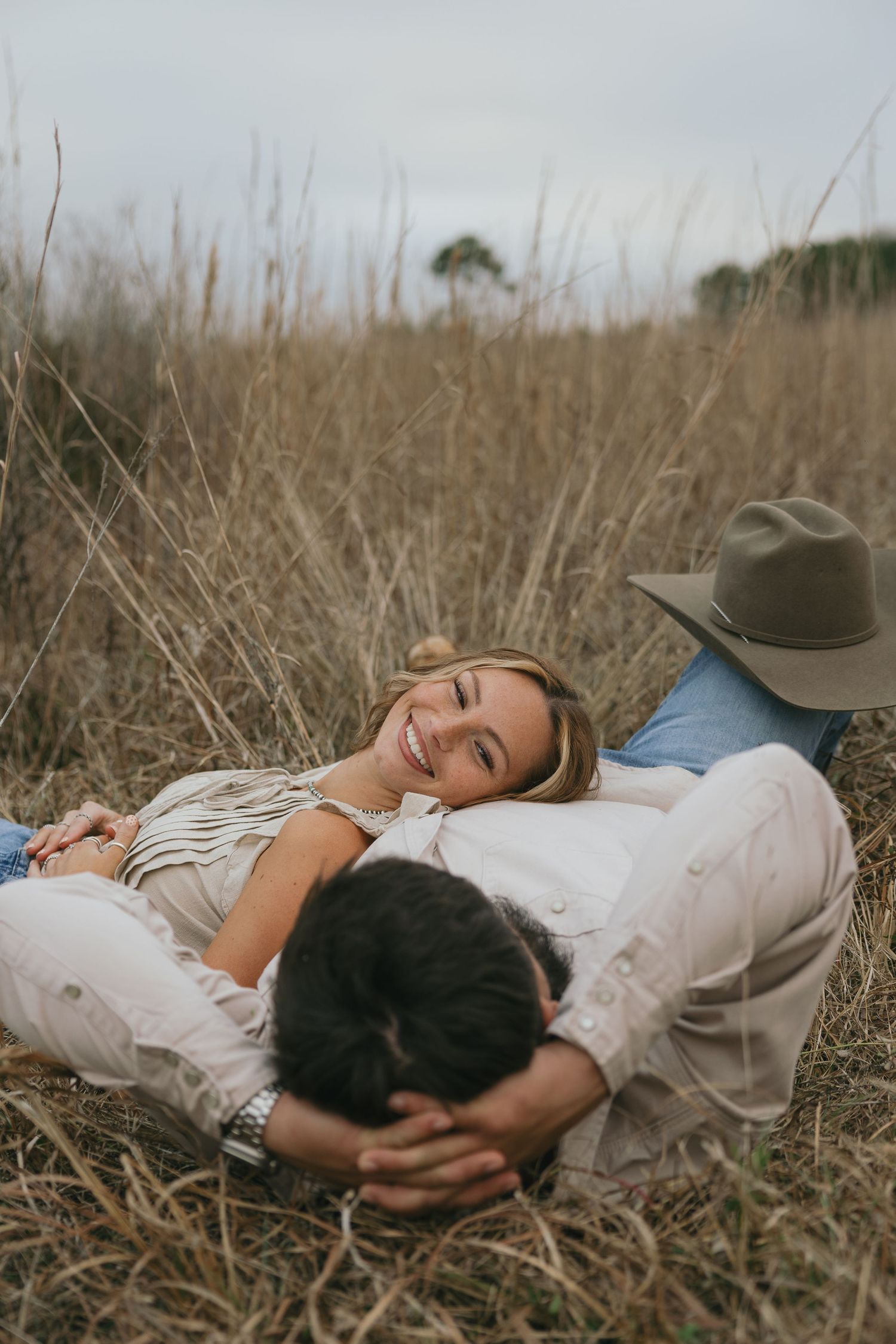 Smiling woman lying in tall golden grass resting on man's chest during romantic outdoor couple session.