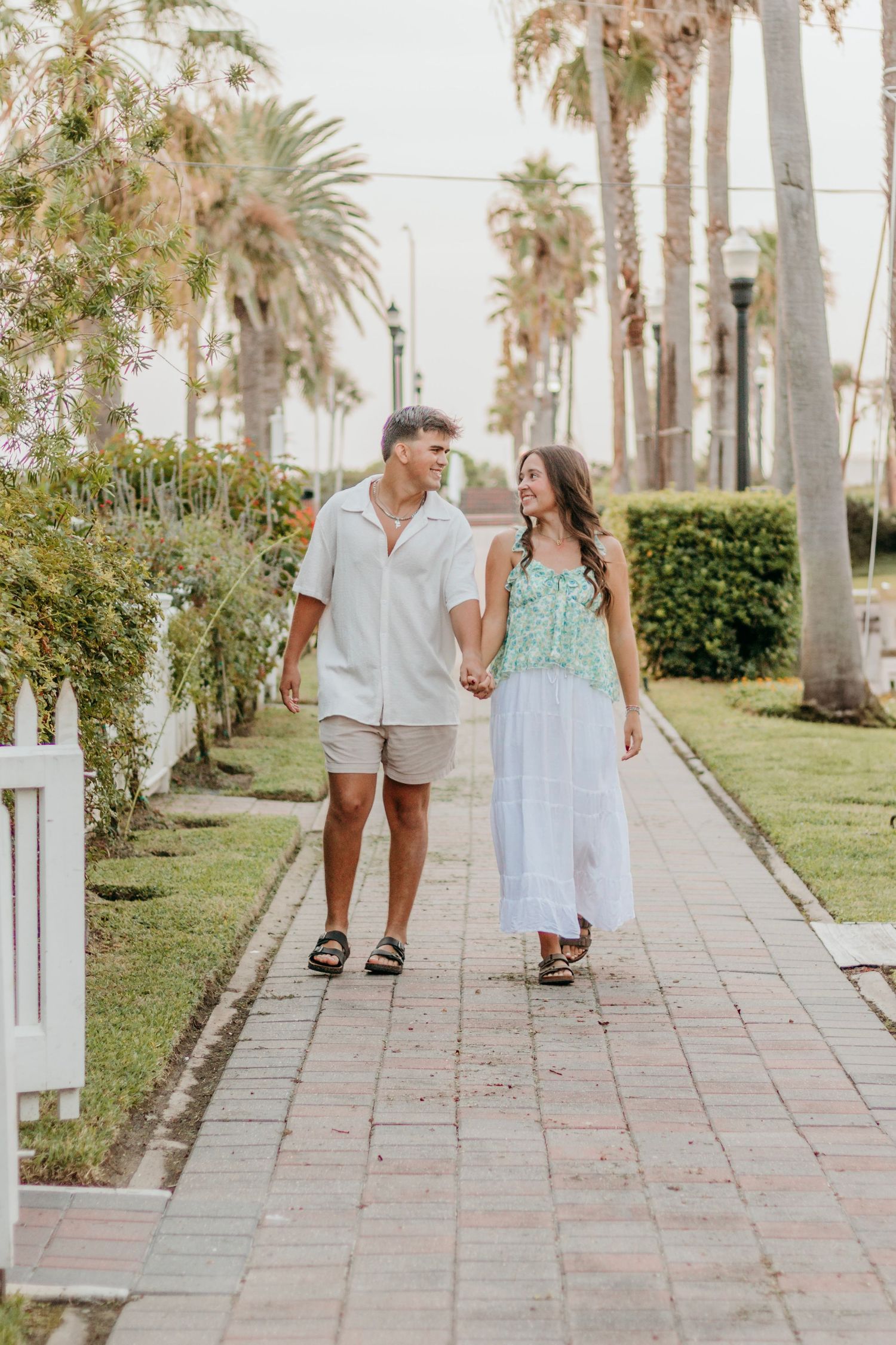 Couple holding hands and walking along a palm tree-lined brick pathway in a tropical setting.