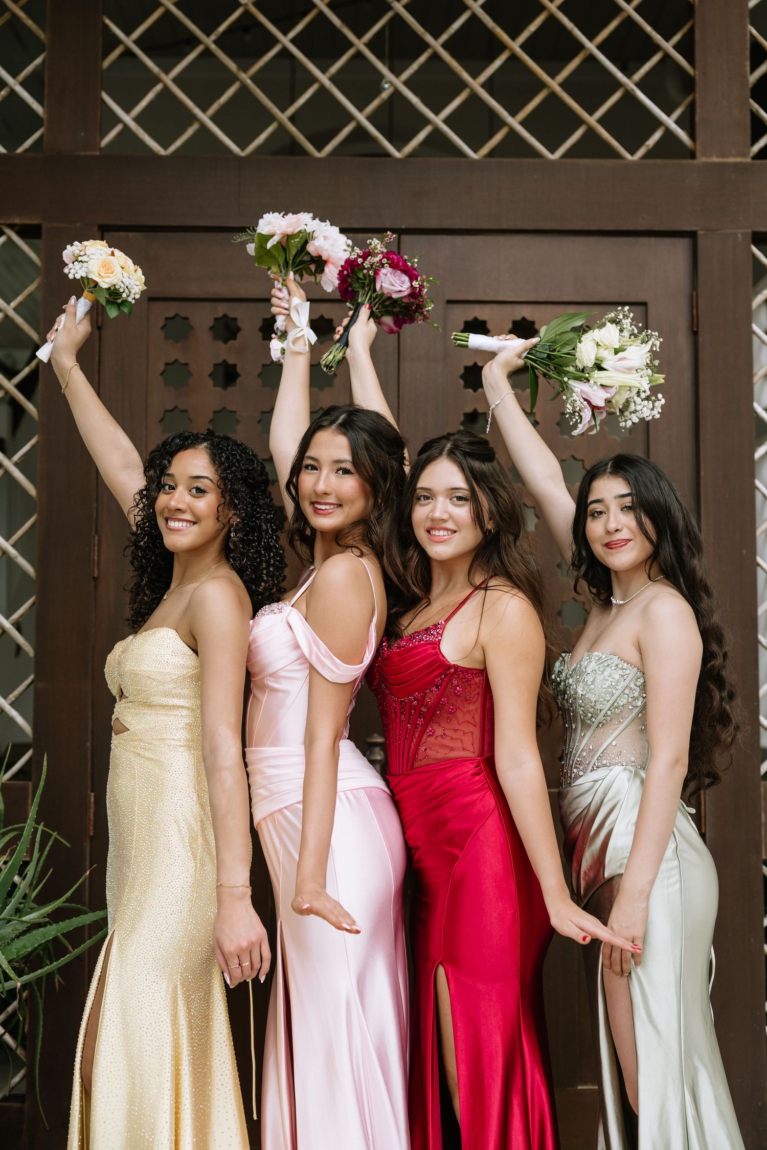 Four young women in elegant prom gowns raising flower bouquets in front of wooden doors.