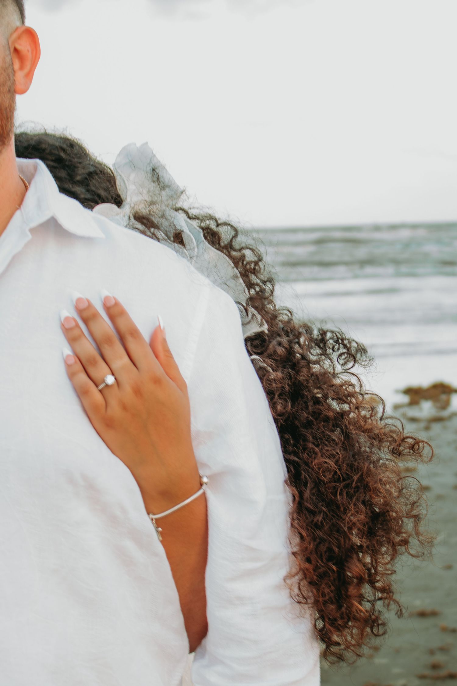 Woman with curly hair placing hand on mans white shirt near ocean, showing ring and bracelet.