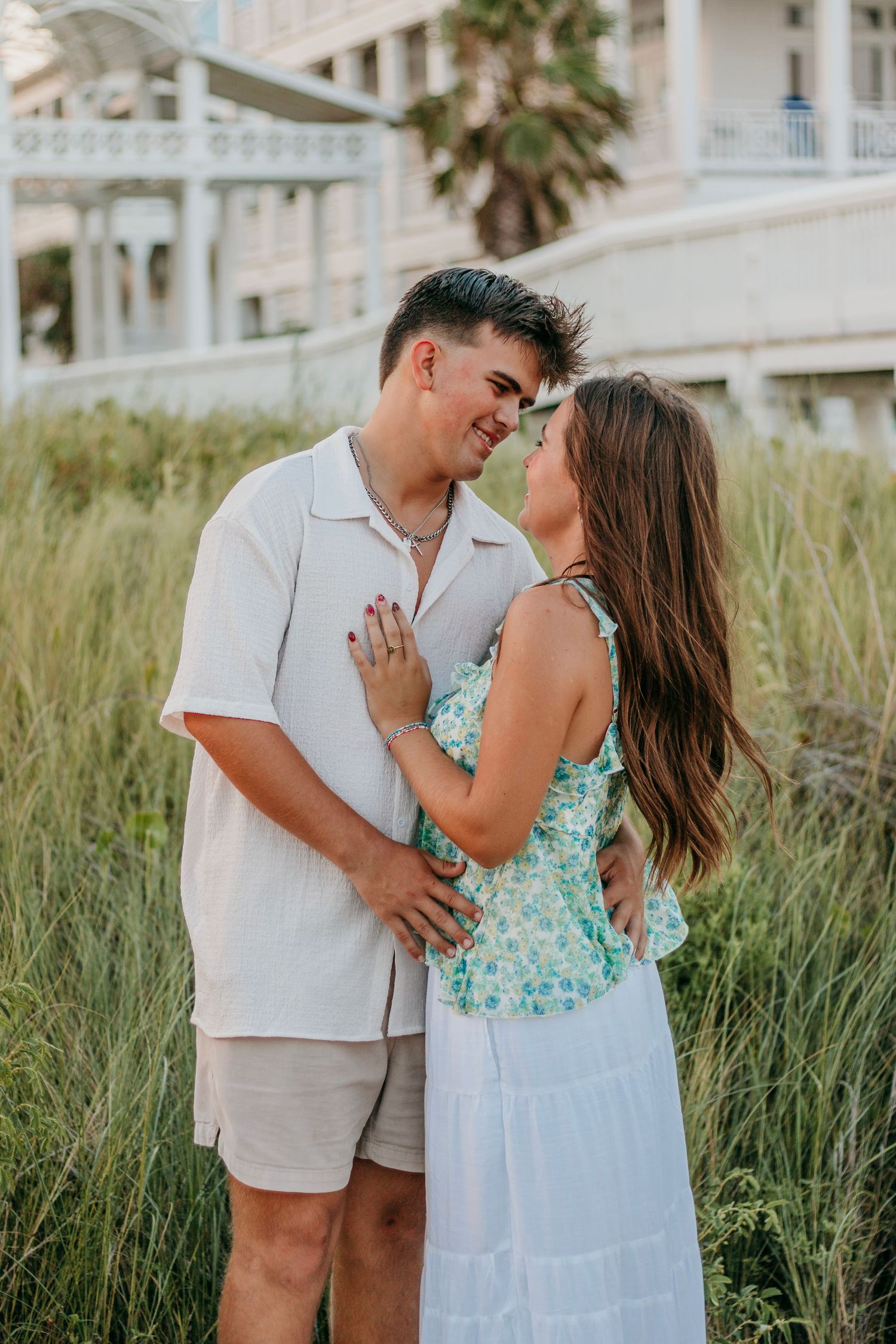 Romantic couple nearly kissing in tall grass near beach resort, woman in floral top and white skirt.