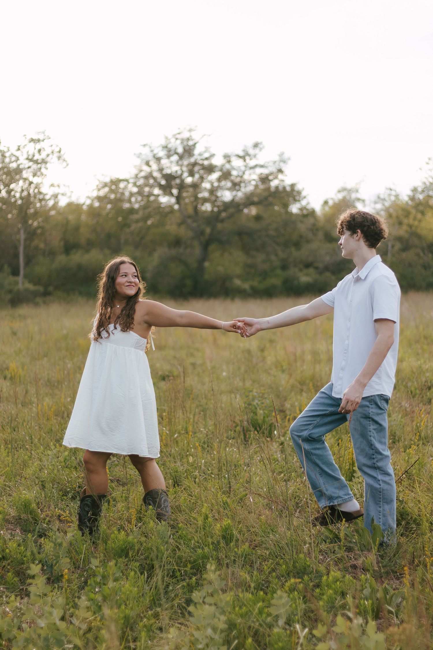Young couple holding hands and dancing in a sunlit meadow, woman in white dress and cowboy boots smiling joyfully.