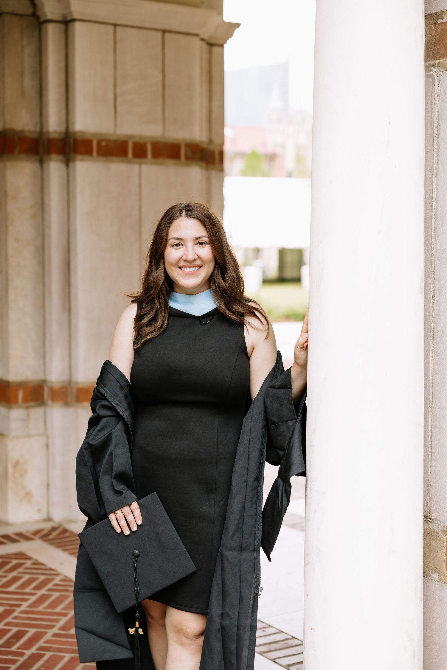 Smiling graduate in black gown holding cap while leaning against a white column.