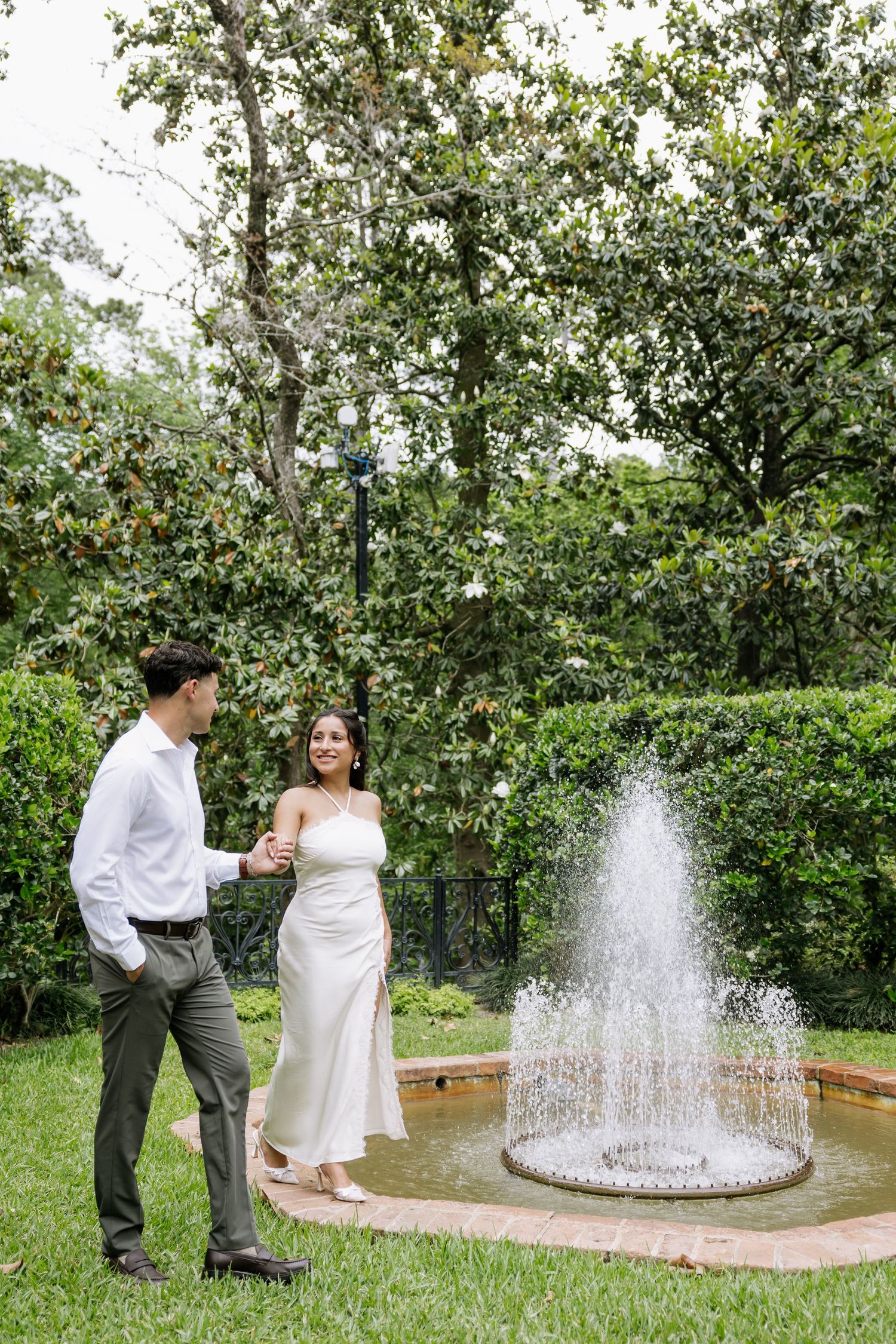 Couple holding hands and smiling near a garden fountain surrounded by lush greenery.