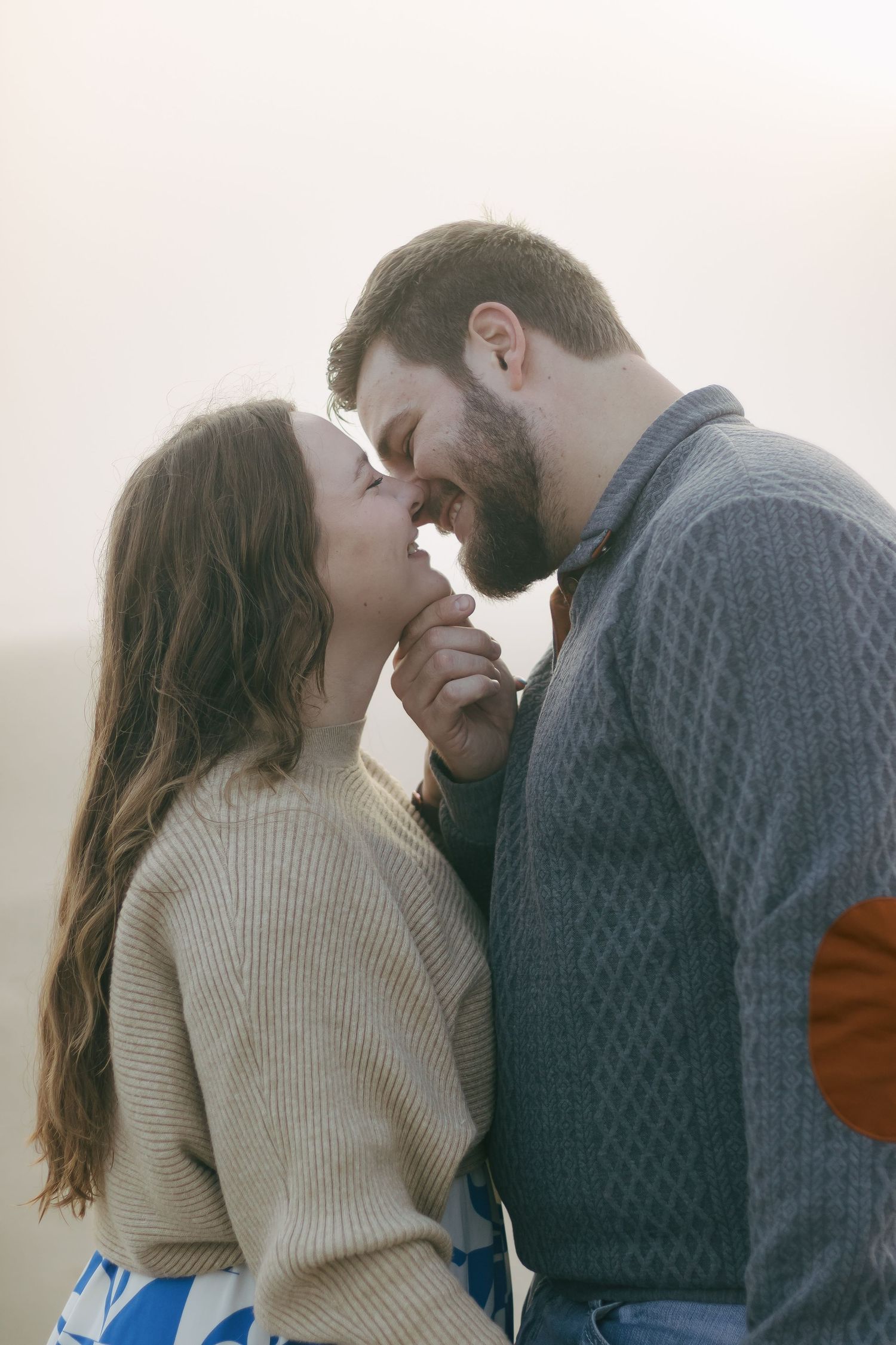 Couple sharing a tender almost-kiss outdoors in soft foggy light, woman in beige sweater holding hands.
