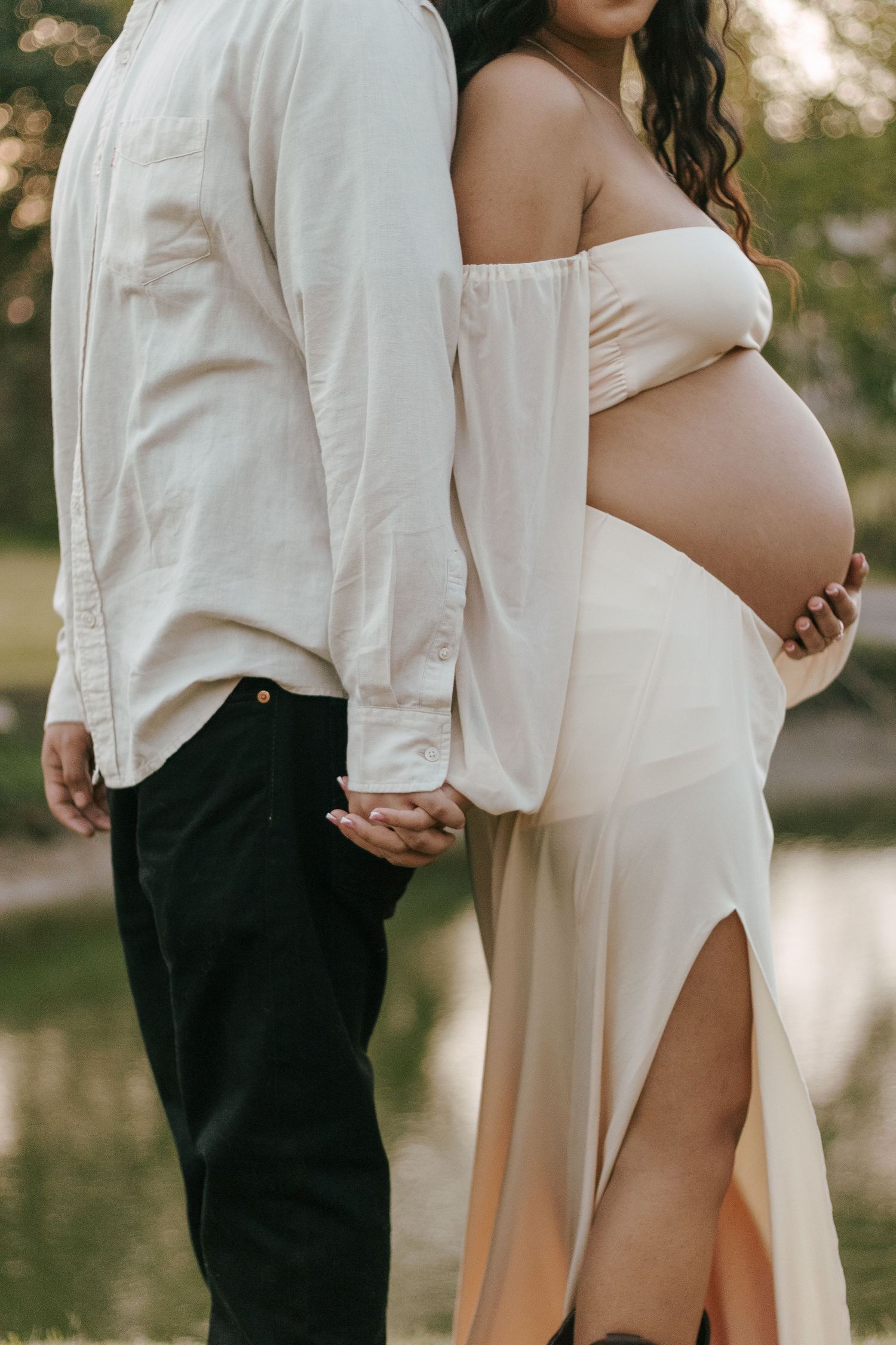 Maternity photo of pregnant woman in cream off-shoulder gown holding bump beside partner near pond.