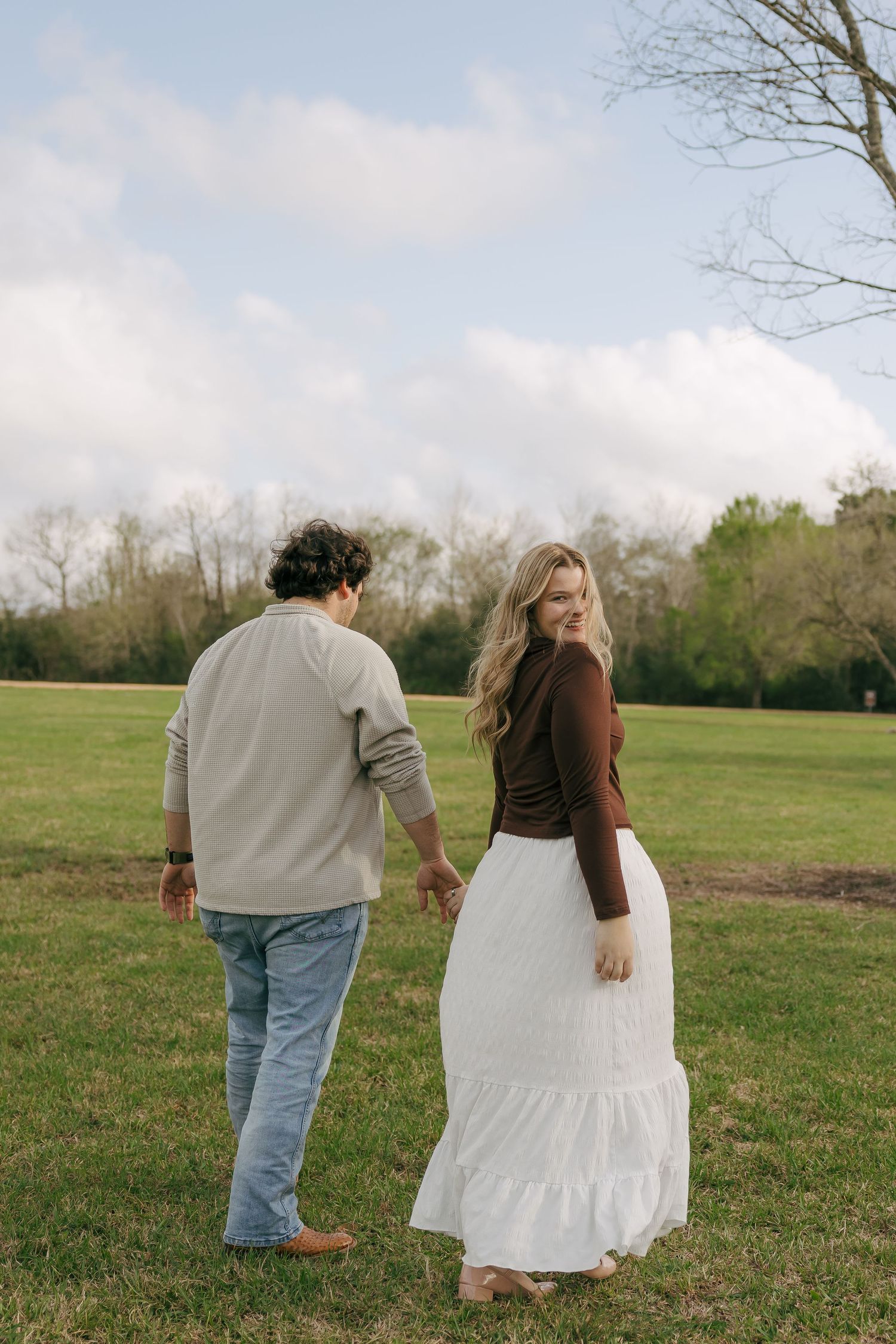 A couple holds hands walking through a green field, the blonde woman in a white skirt glances back smiling.