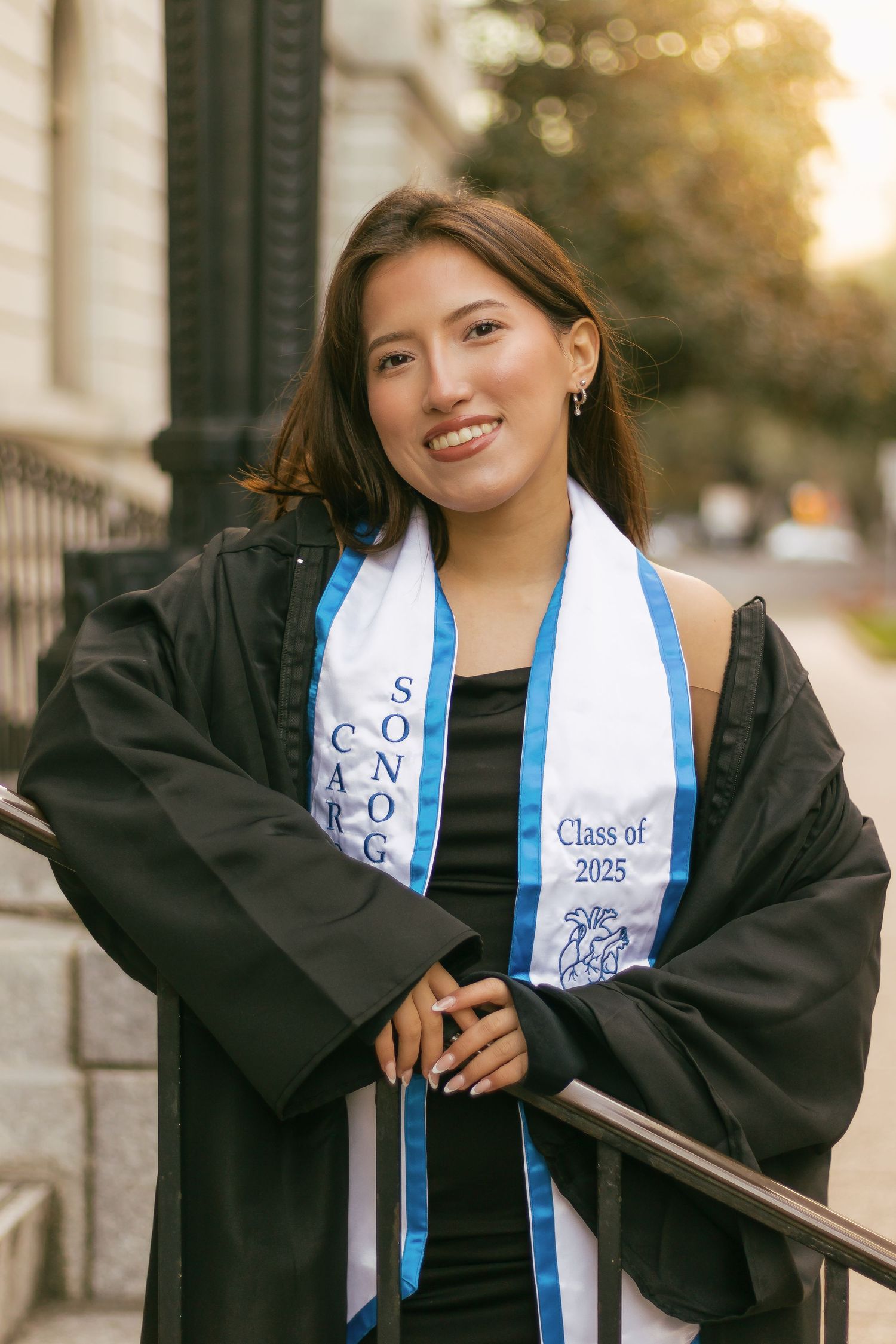 Smiling graduate in black gown and Class of 2025 stole posing outdoors at golden hour.