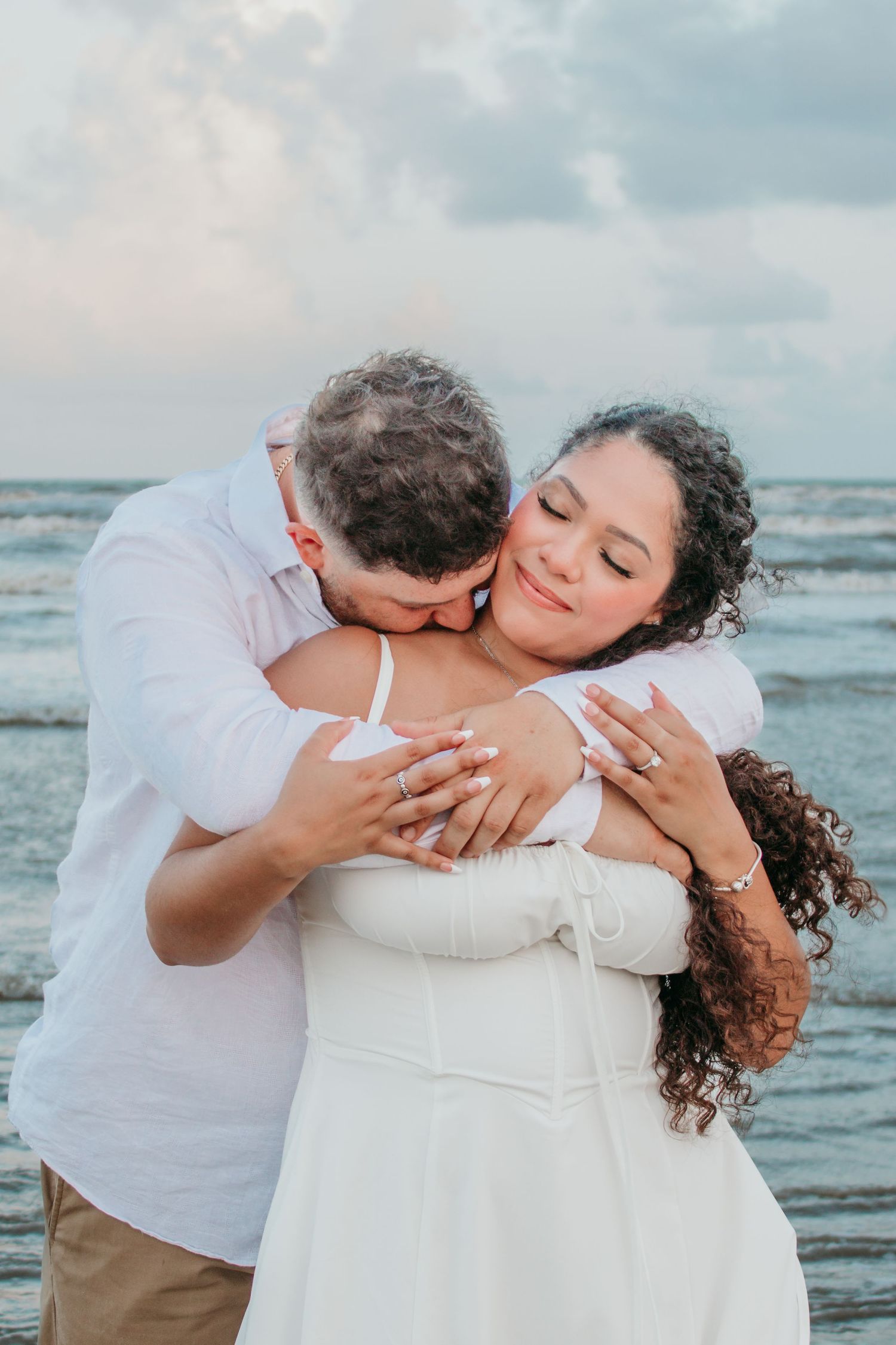 Couple sharing a tender embrace on the beach at sunset, woman in white dress with eyes closed peacefully.