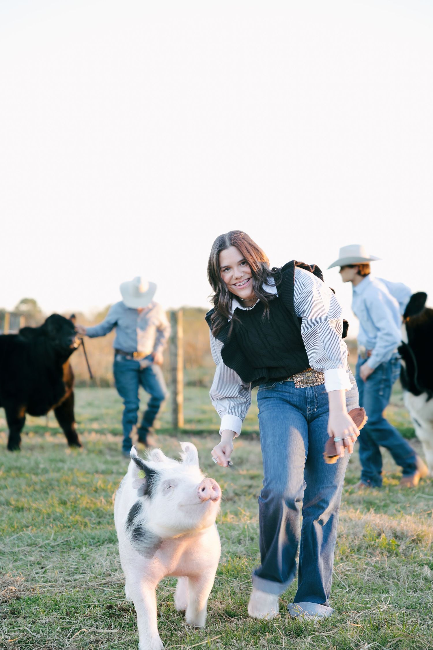 A young woman in western attire smiles while interacting with a spotted piglet on a farm with cattle in the background.