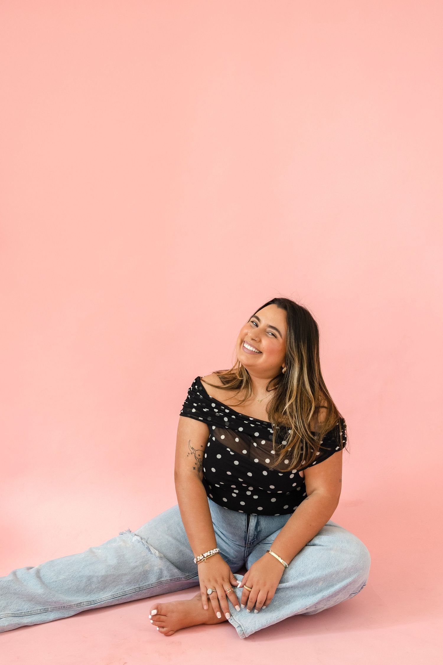 Happy woman in polka dot off-shoulder top sitting on pink floor looking up with a smile.