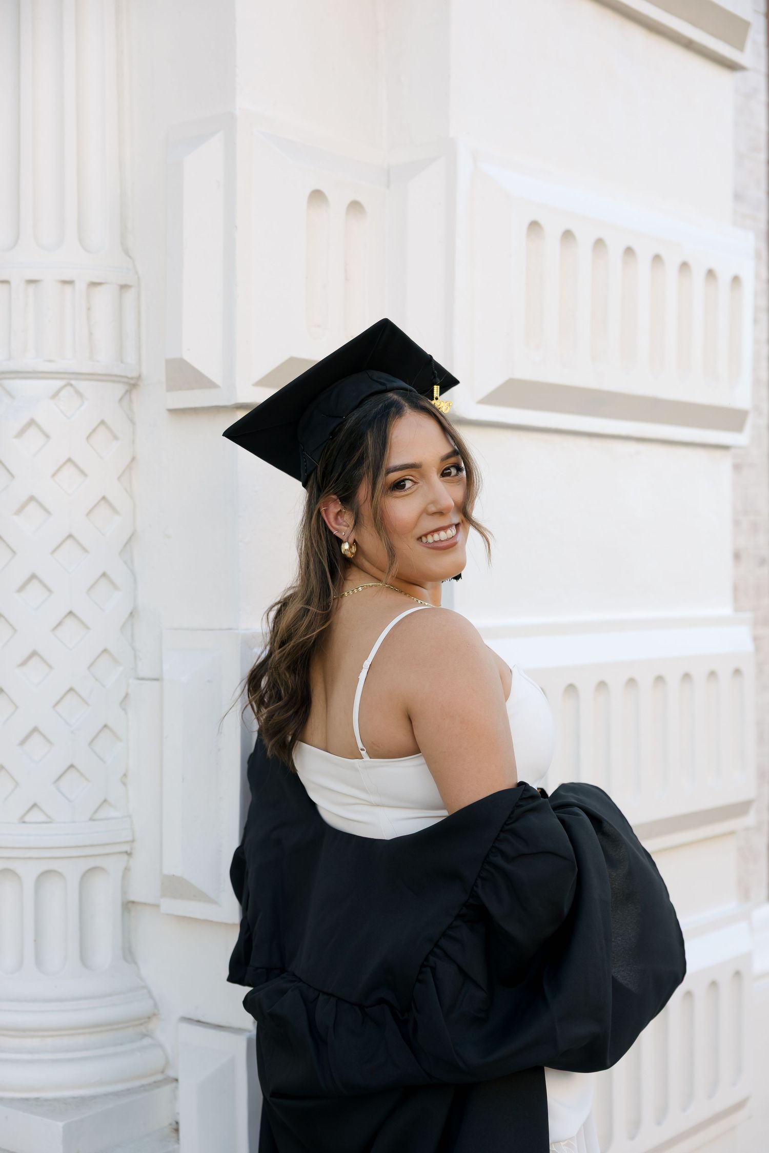 Smiling young woman in black graduation cap and gown posing against a white architectural wall.