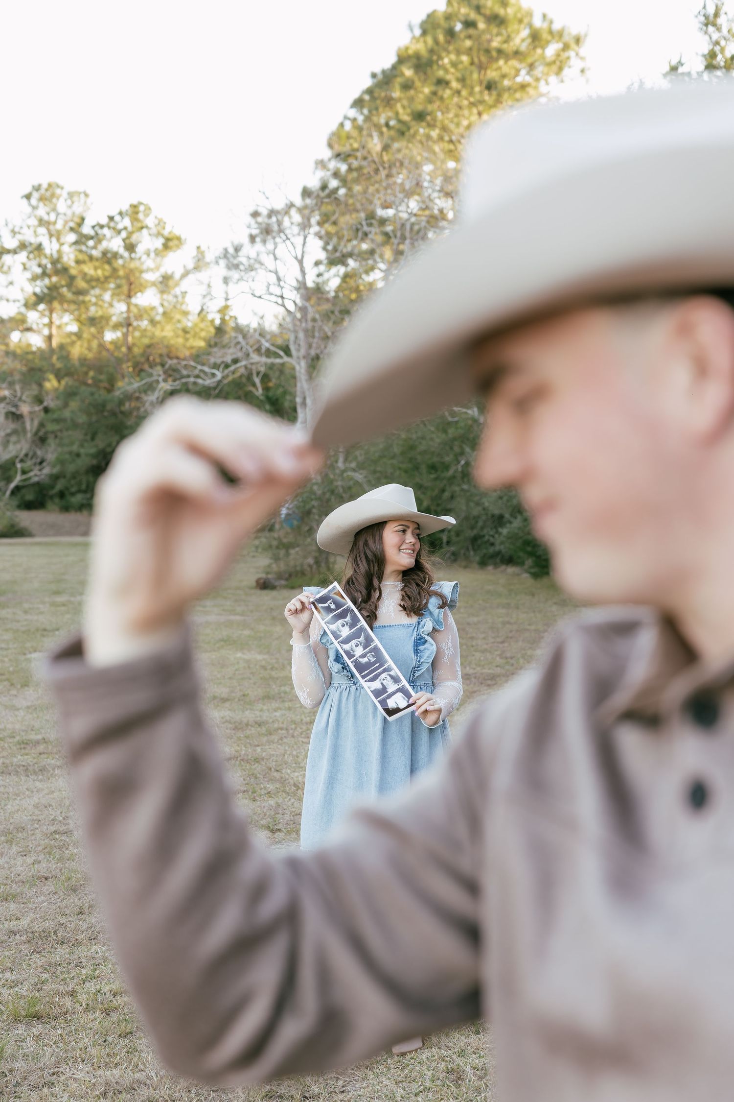 Woman in blue dress with engagement sash smiles as man in cowboy hat tips his hat in a field.