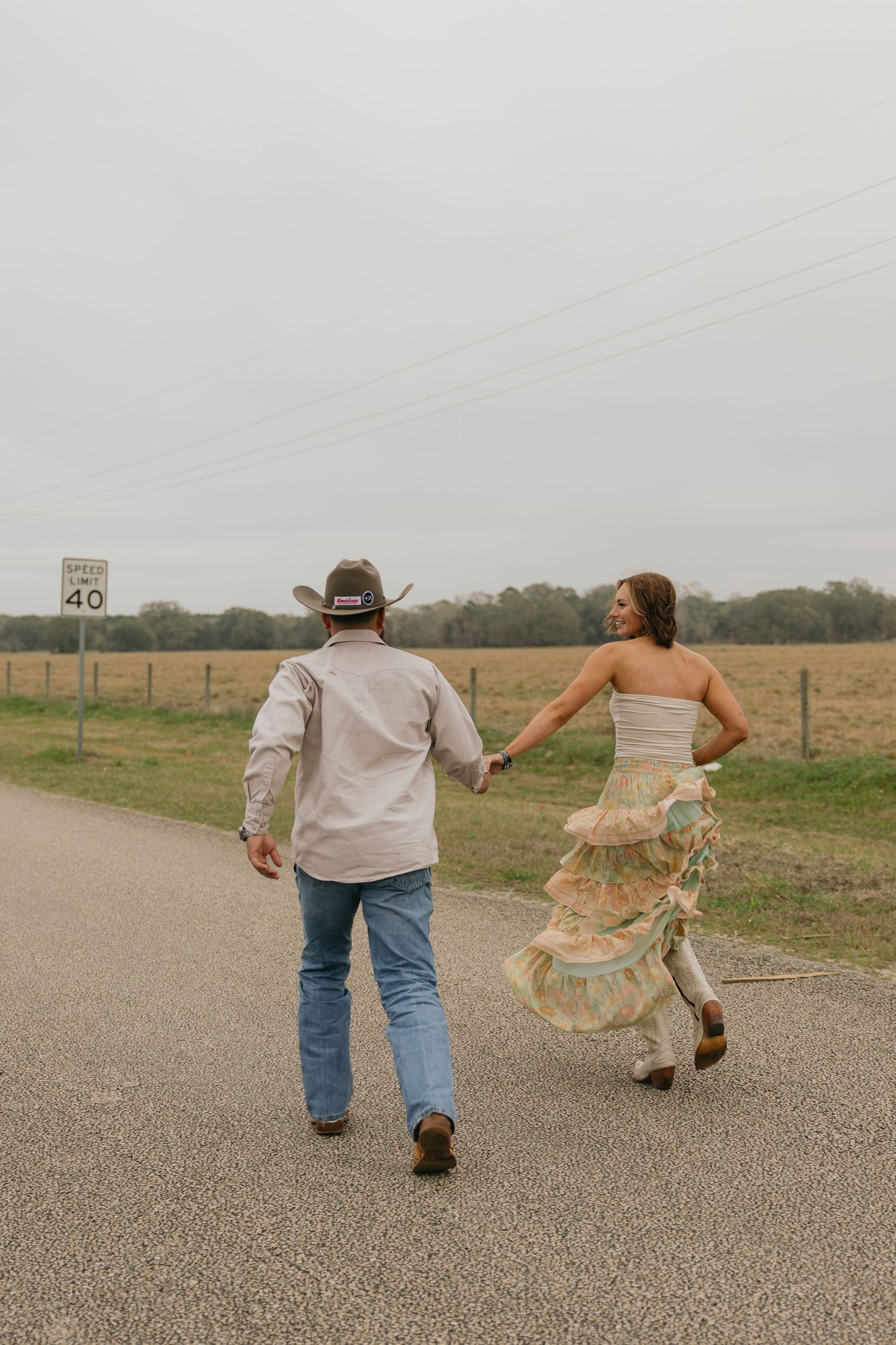 A couple walks hand-in-hand down a rural road, the woman in a ruffled dress and cowboy boots beside a speed limit sign.
