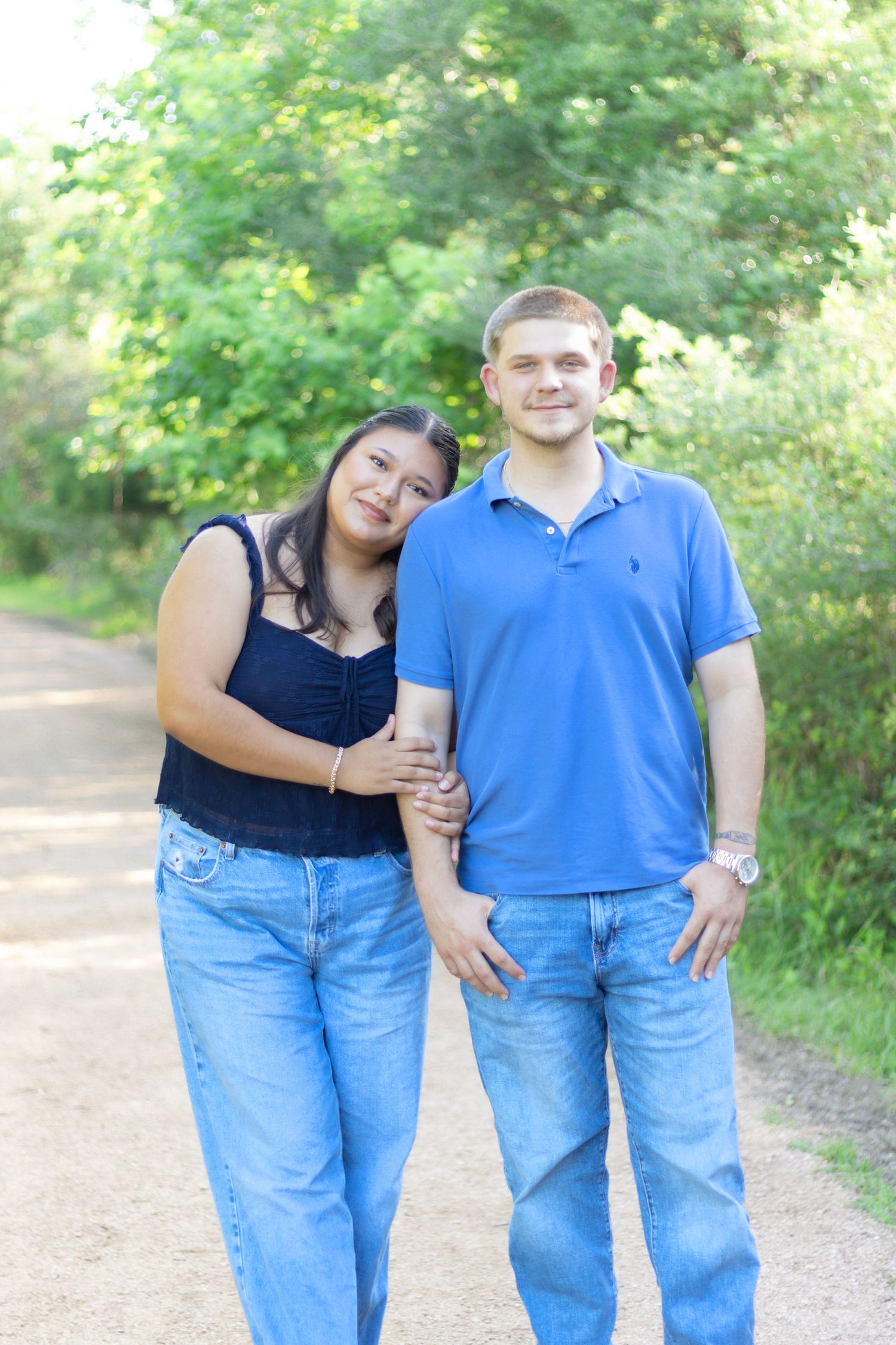 Young interracial couple standing together on a tree-lined path, woman in navy top leaning on mans arm.