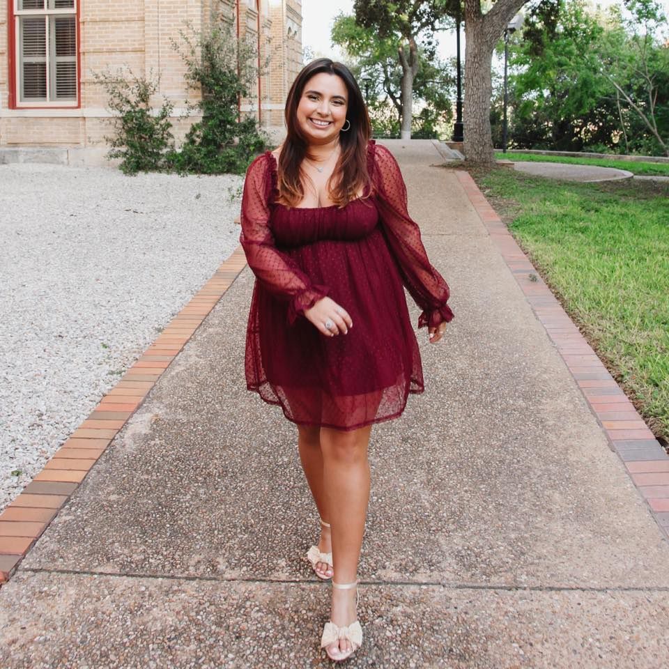 Young woman in burgundy long-sleeve dress walking on brick pathway surrounded by green trees.