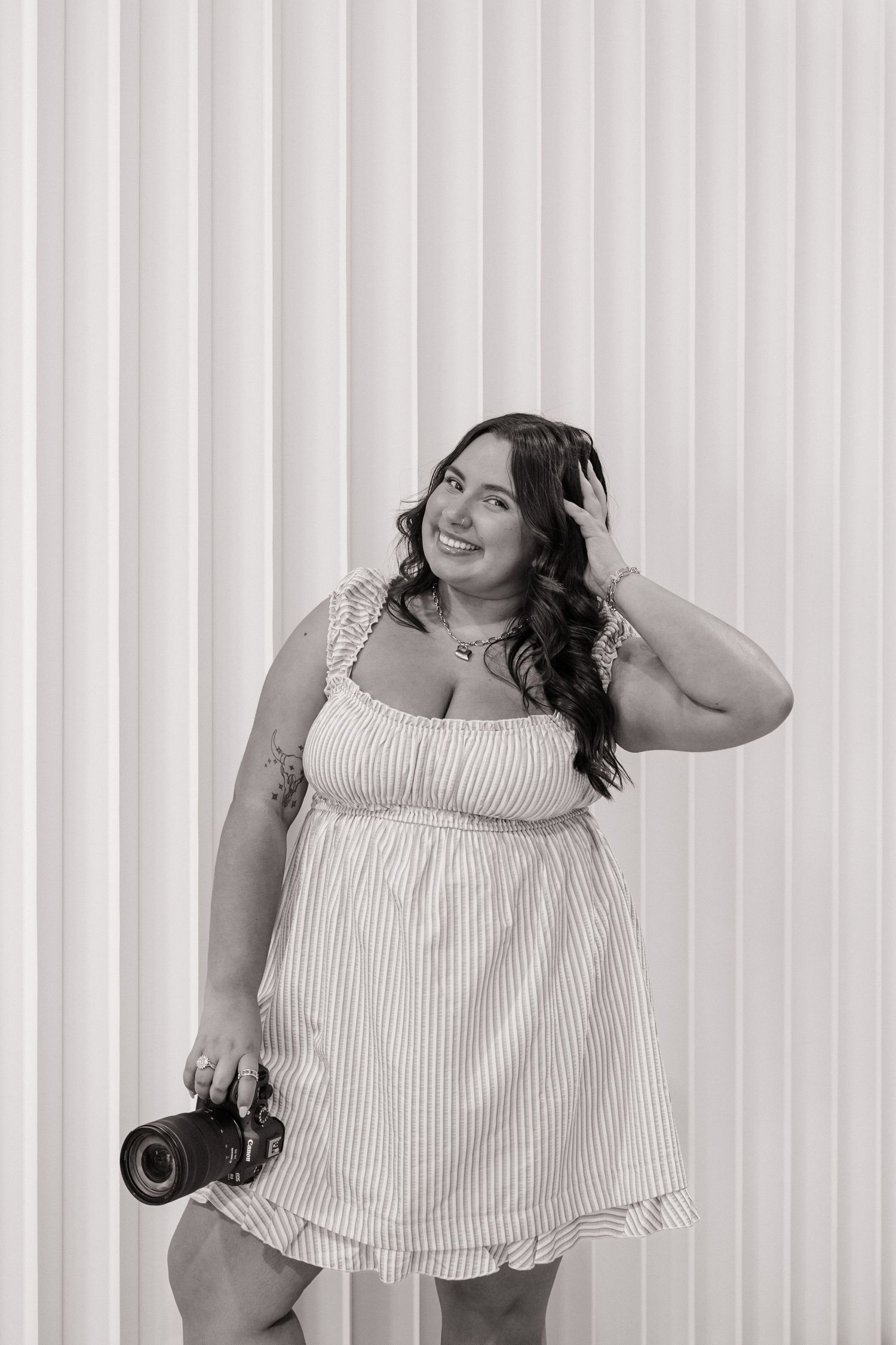 Black and white portrait of a smiling woman holding a camera against a white wall.