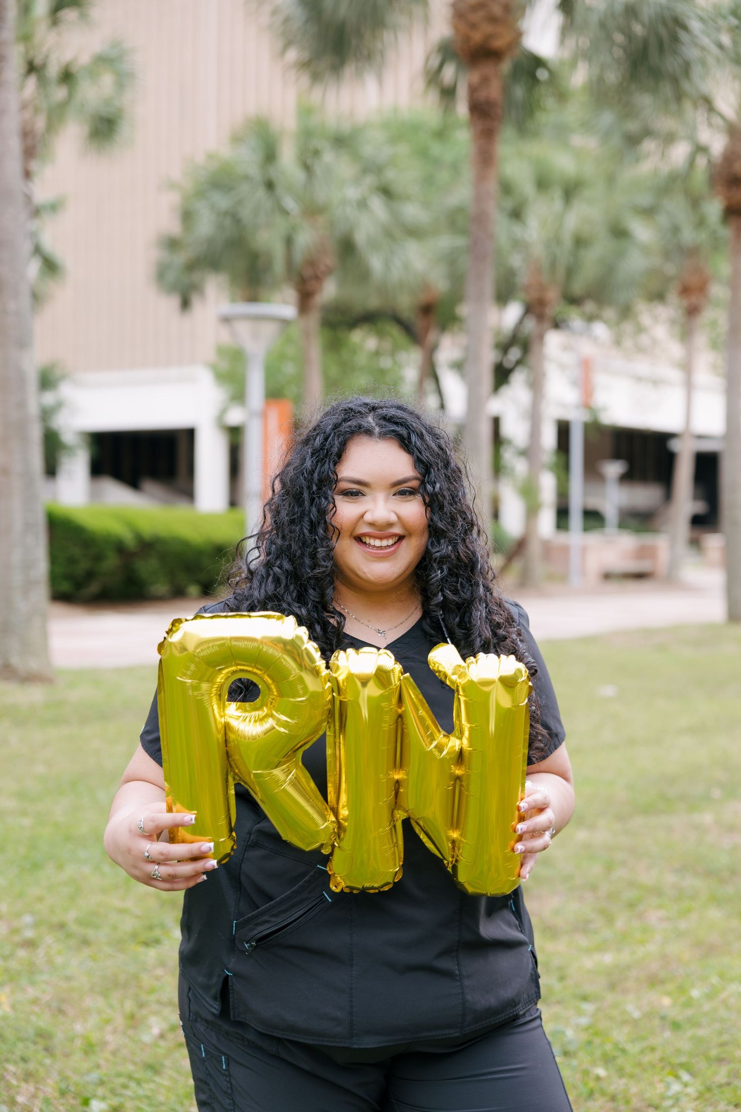 Smiling nurse in black scrubs holding gold RN letter balloons outdoors near palm trees.
