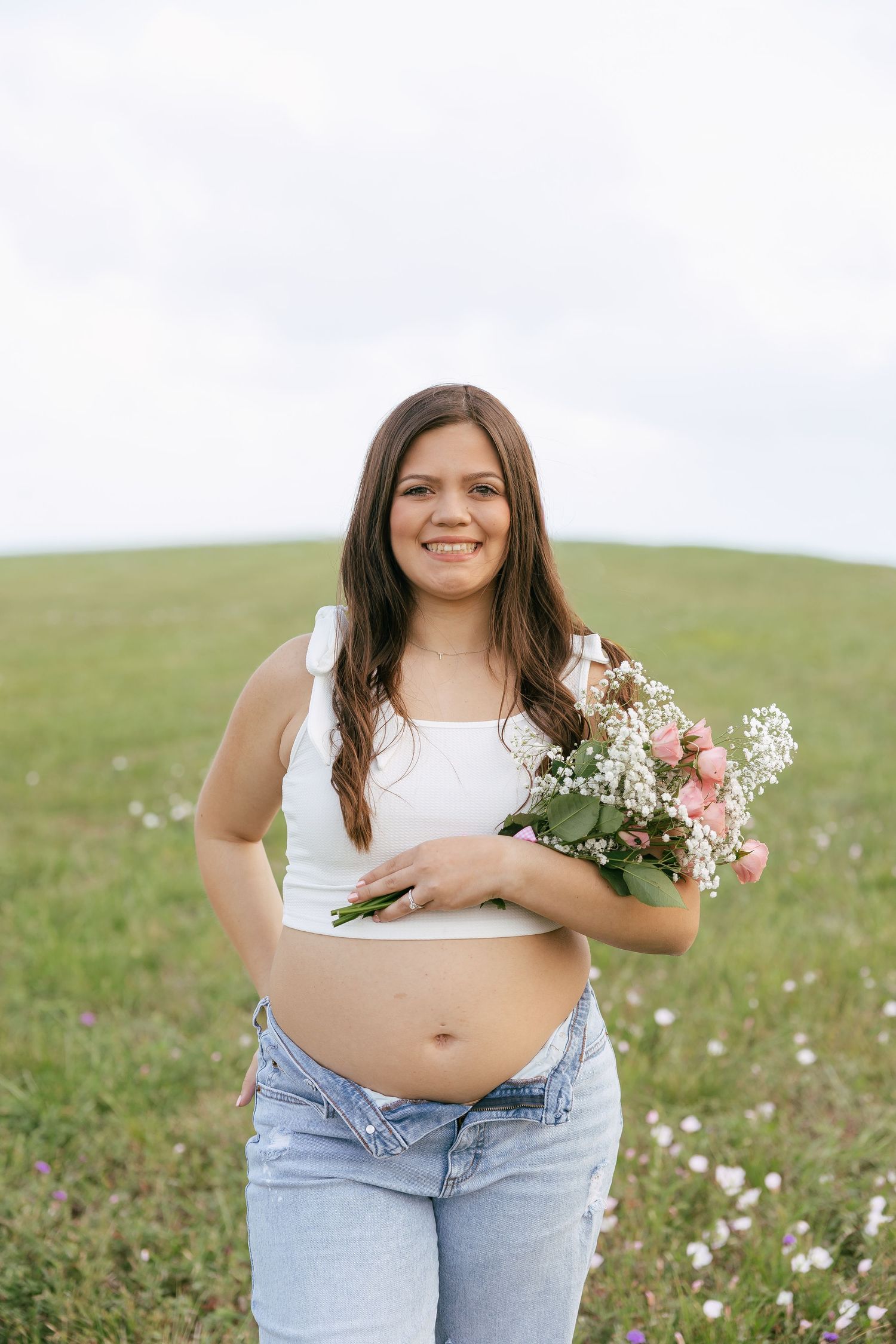 Smiling pregnant woman in white top and jeans holding a flower bouquet in a green field.