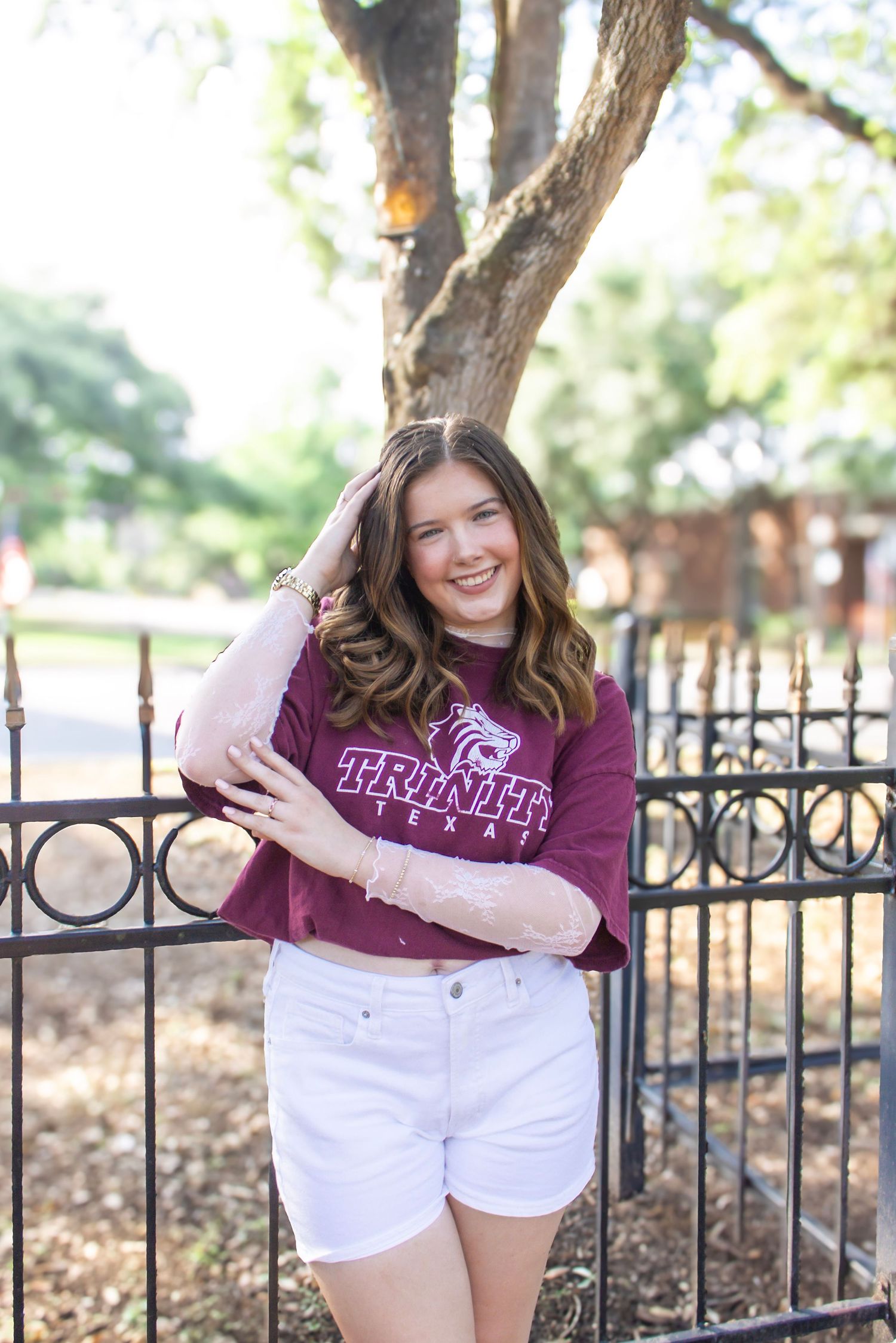 Smiling young woman in Trinity University shirt and white shorts leaning on iron fence outdoors.