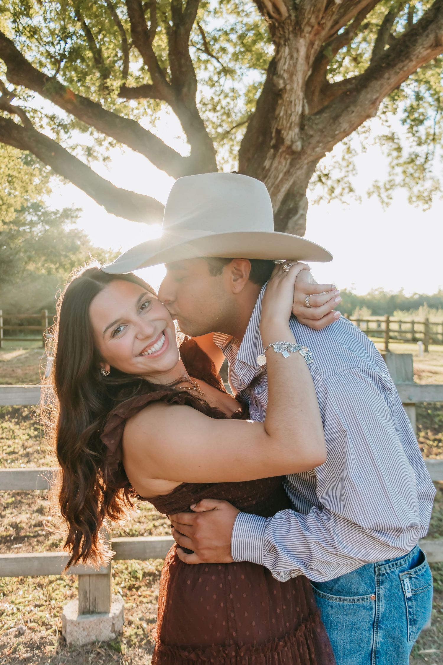 Couple embracing under a large oak tree at golden hour, man in cowboy hat kissing womans cheek outdoors.
