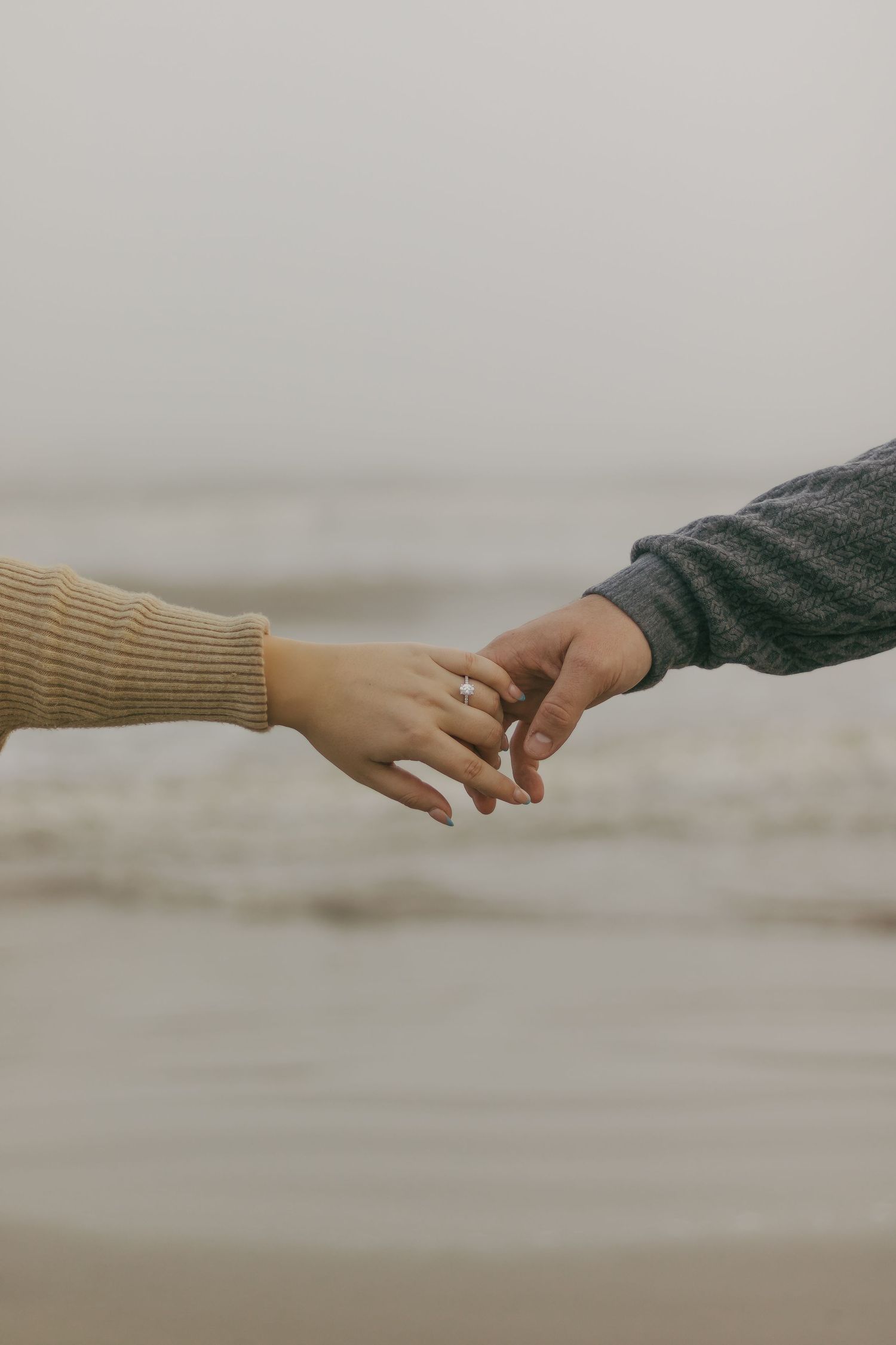 Close-up of couple gently touching fingers on foggy beach, woman wearing engagement ring.