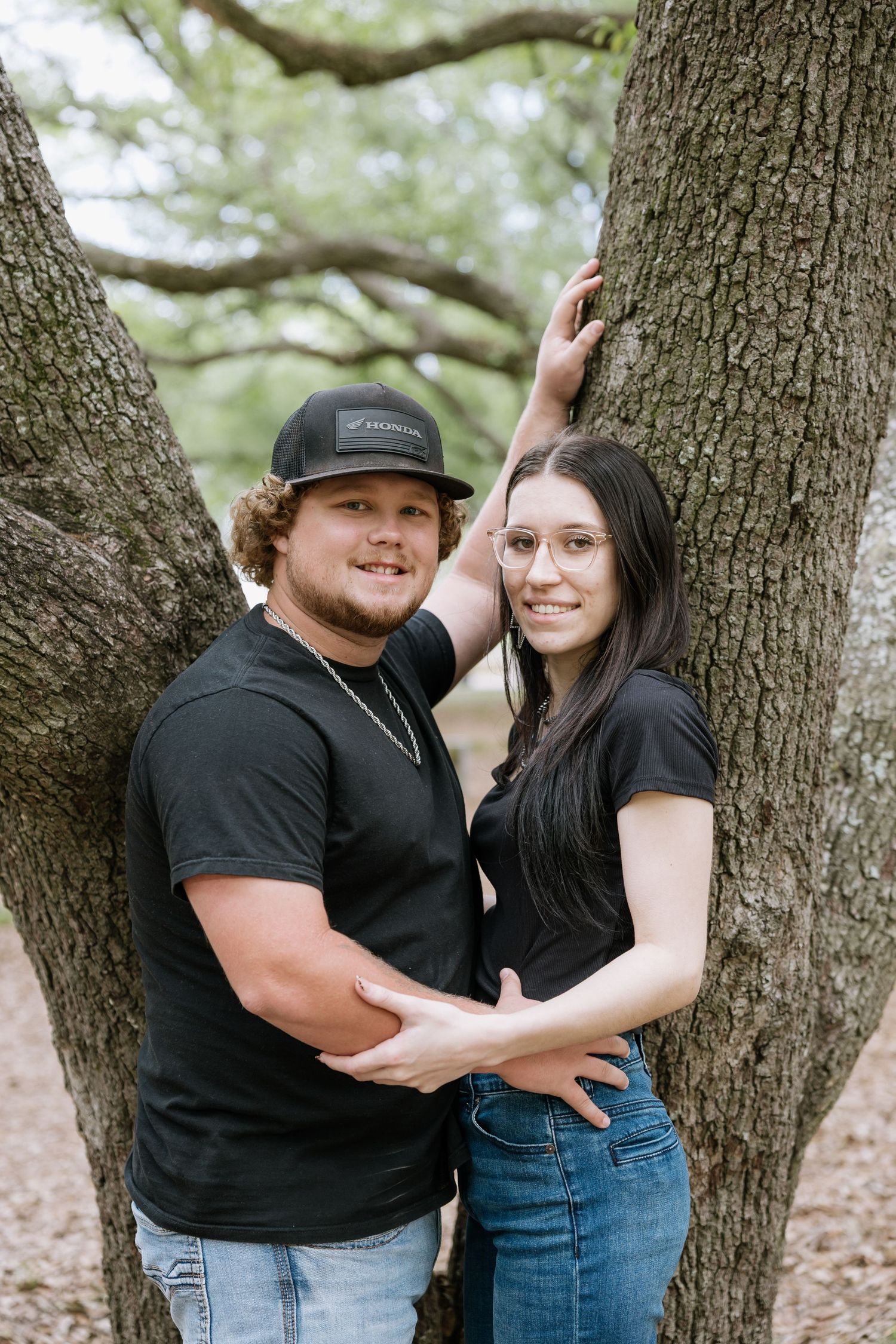 Couple in matching black outfits smiling together between two trees in a wooded setting.
