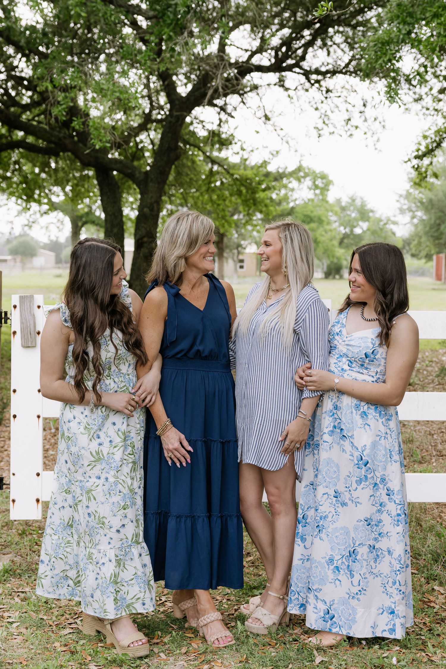 Four women in blue and floral dresses smiling together near a white fence outdoors.